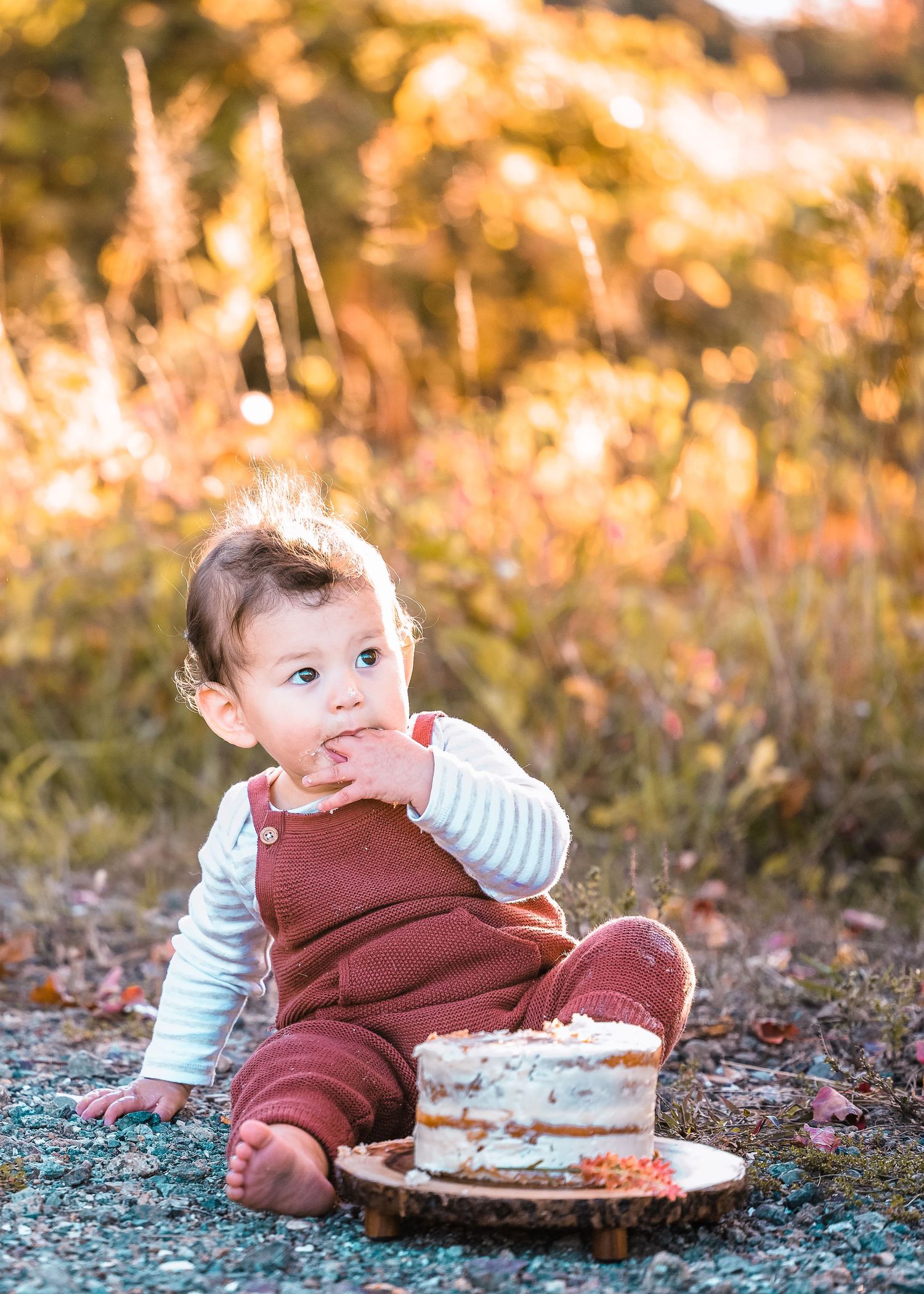 Baby in burgundy overalls sits outdoors with cake enjoying a sweet first birthday celebration.