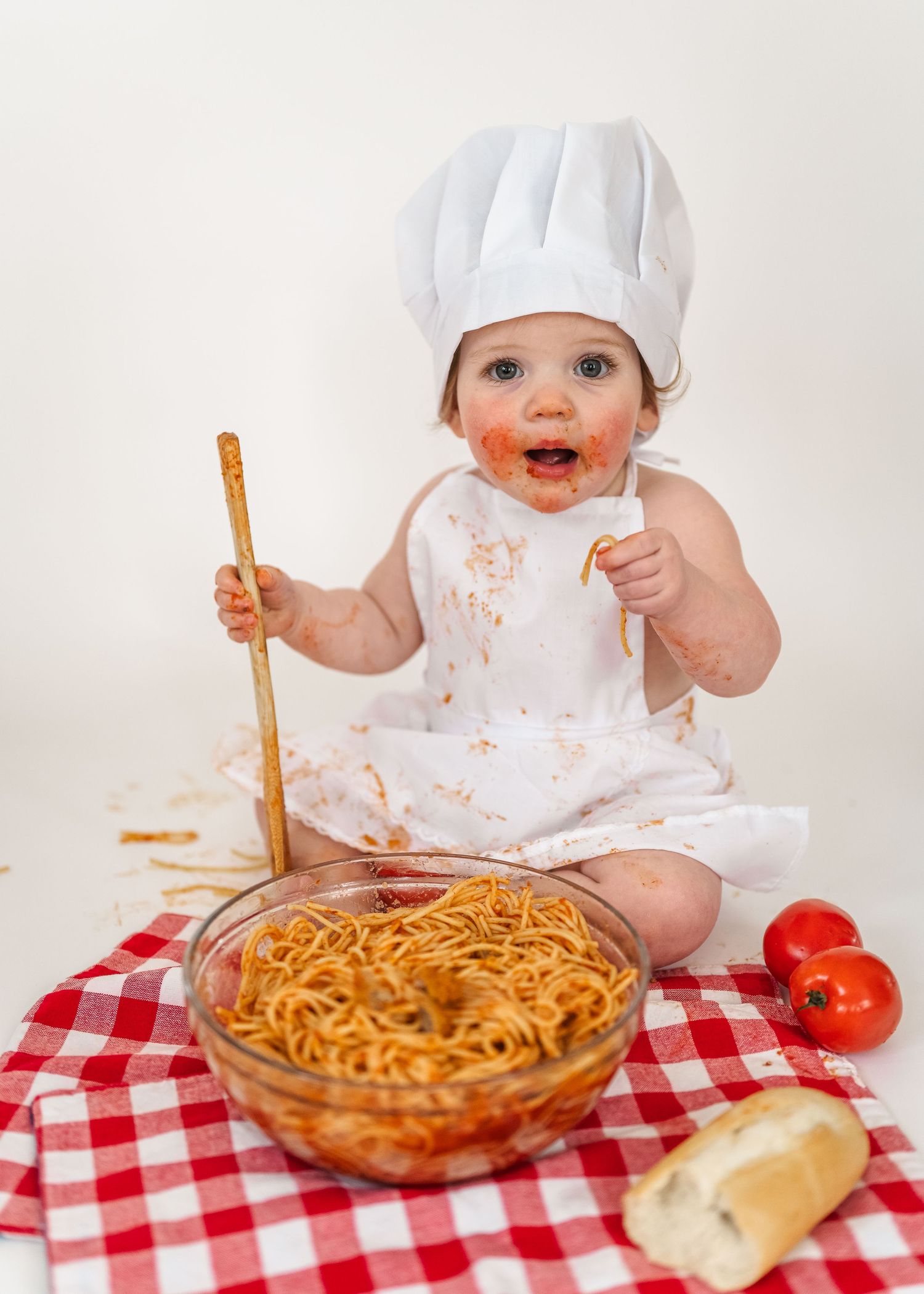 Spaghetti Smash first birthday. A baby in a chef's hat sits at a table with spaghetti and tomatoes on a red checkered tablecloth.