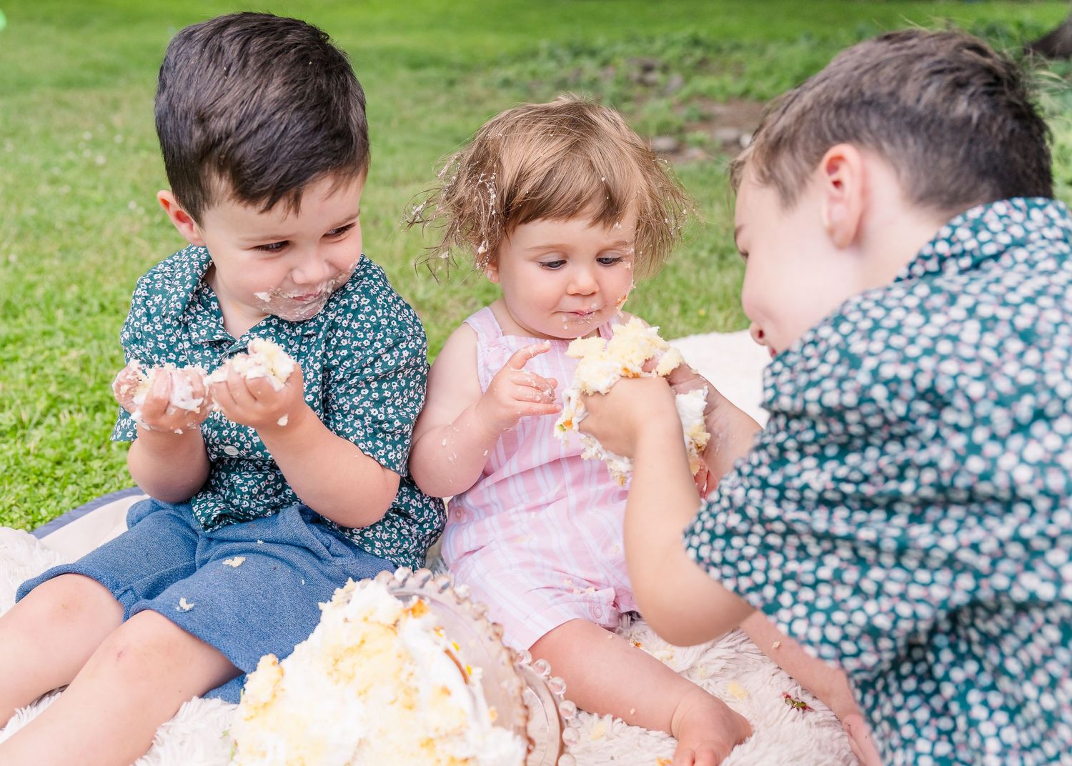 Three children sitting on blanket outdoors sharing birthday cake together.