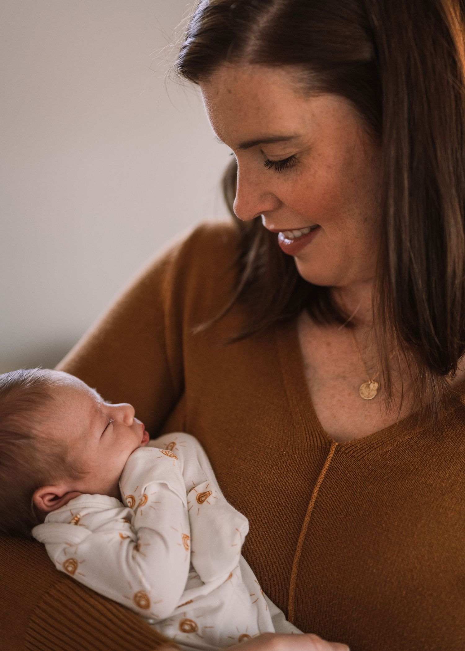 Mother in brown sweater gazes lovingly at sleeping newborn in white onesie.