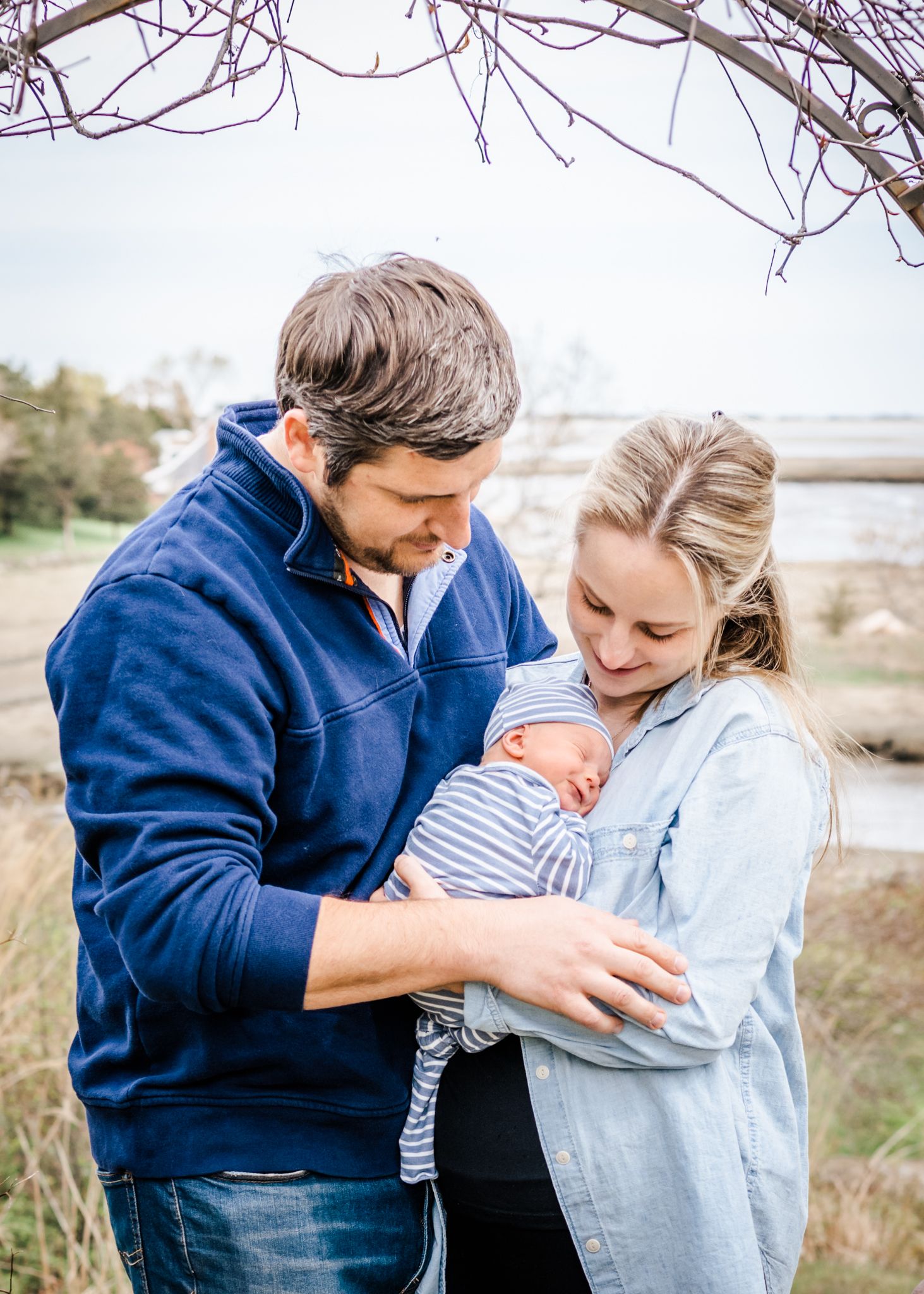 A couple embracing their newborn baby outdoors during an autumn day with bare branches visible.