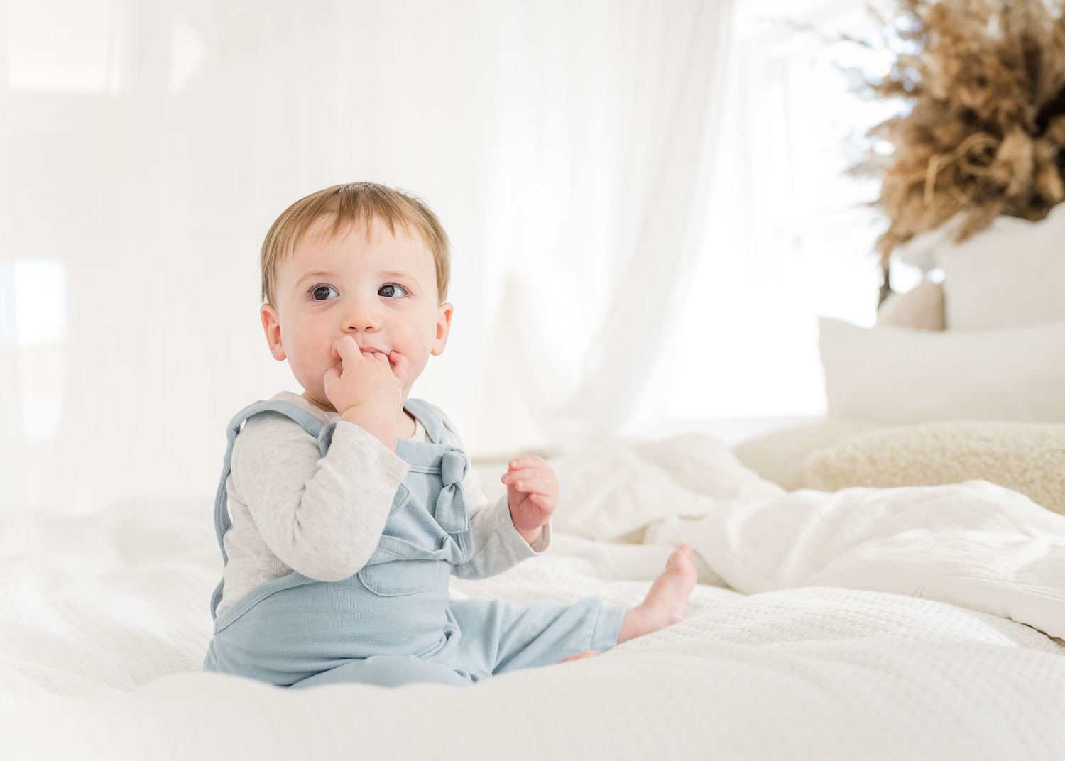 Baby wearing light blue pajamas sits on white bedding while touching their mouth with one hand.