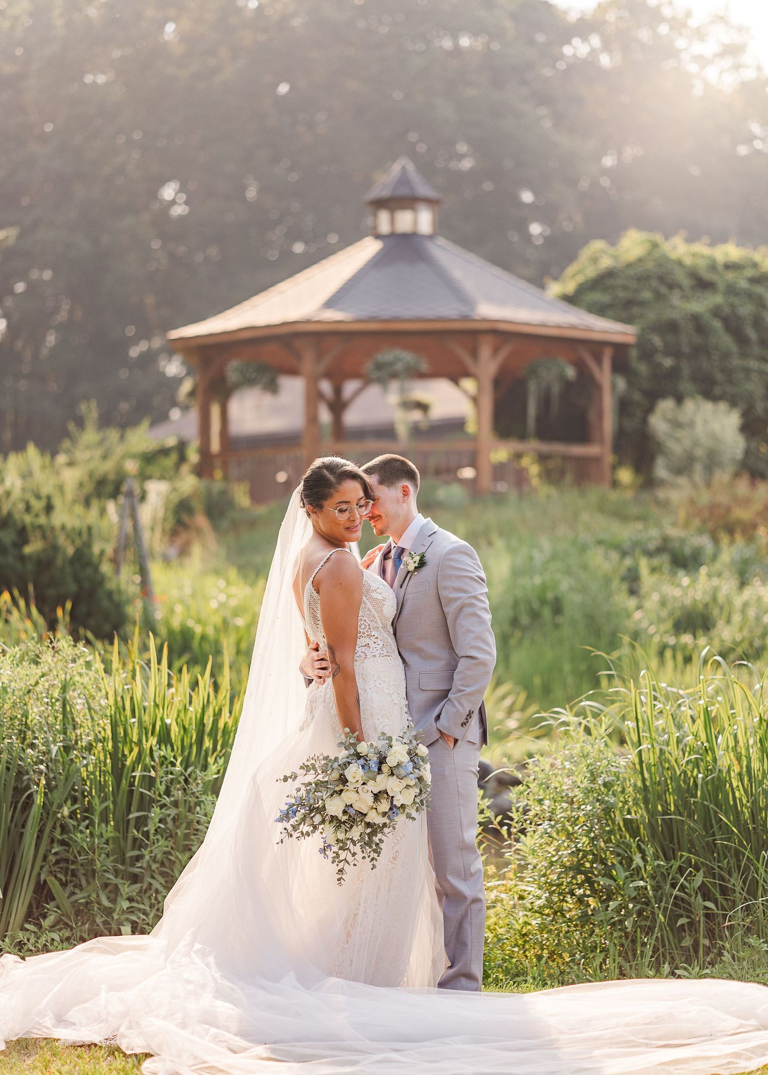 A romantic wedding couple poses in front of a wooden gazebo at sunset in a garden filled with wildflowers.