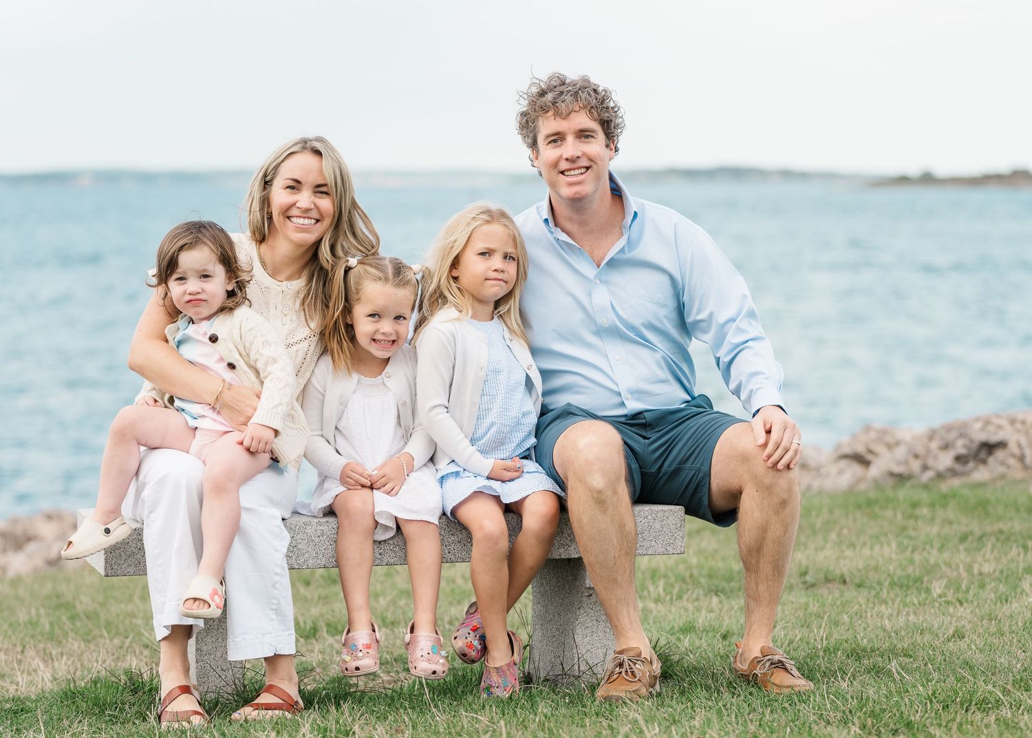 A family sits together on a grassy hill overlooking the ocean in coordinating white and blue outfits.