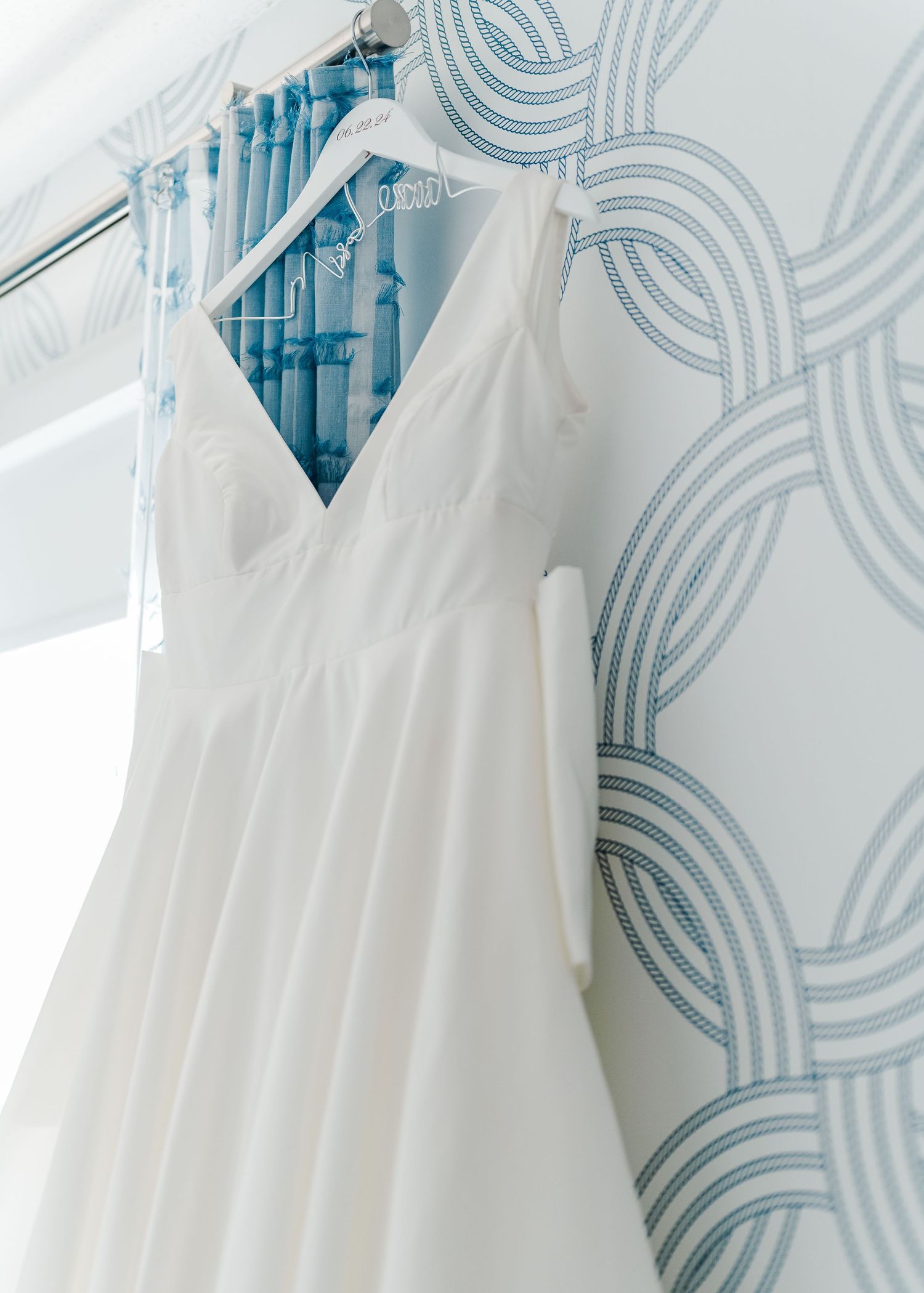 Elegant white wedding dress hanging against blue and white wallpaper in natural light.