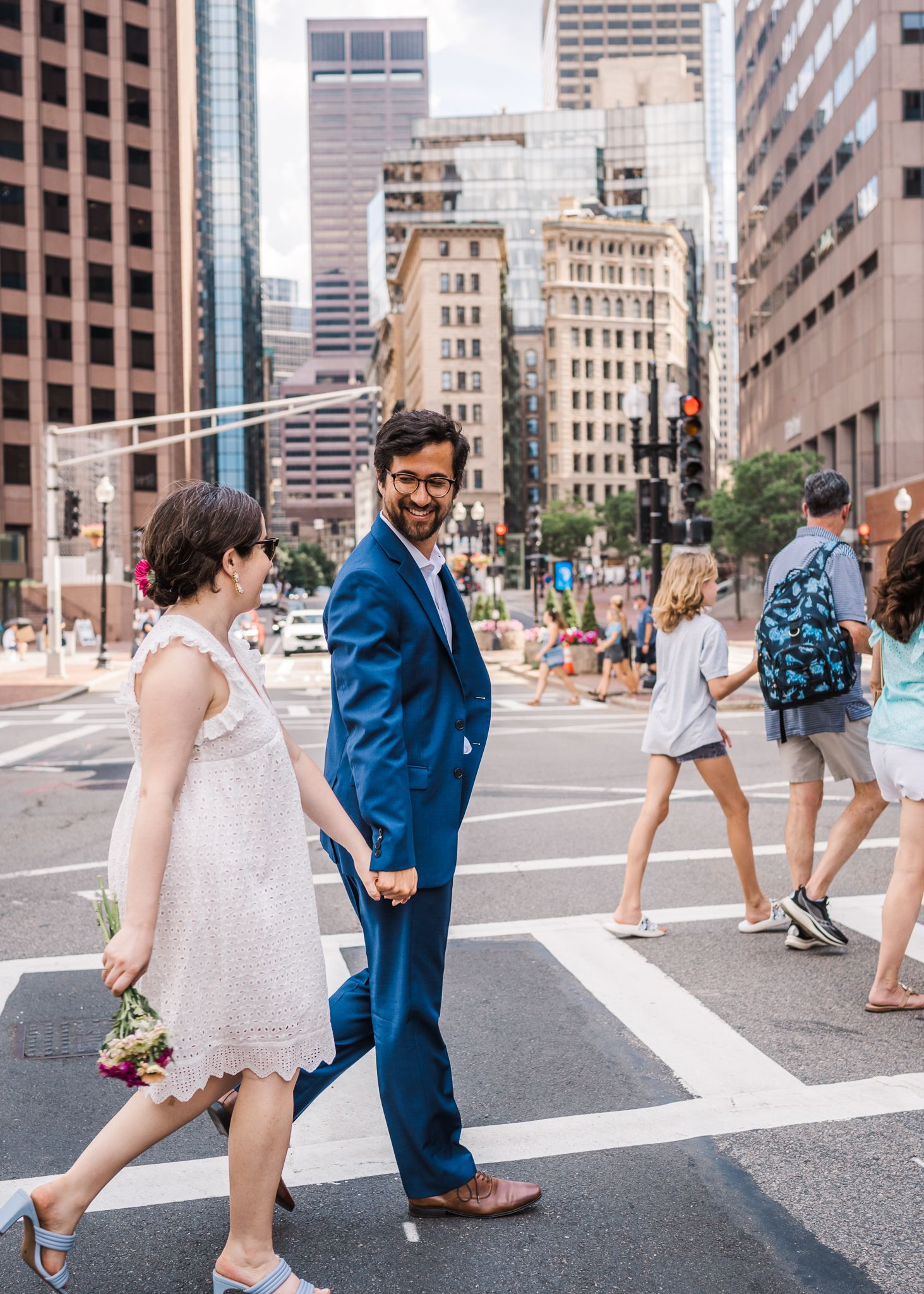 A couple walking across an urban crosswalk with downtown buildings in the background.