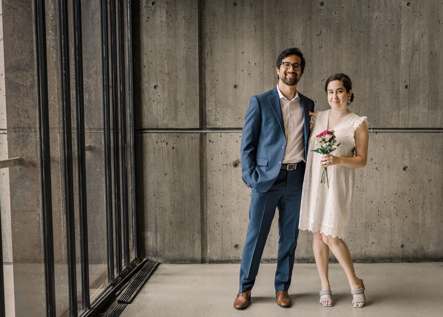 Newlyweds stand together against concrete wall in modern architectural setting.