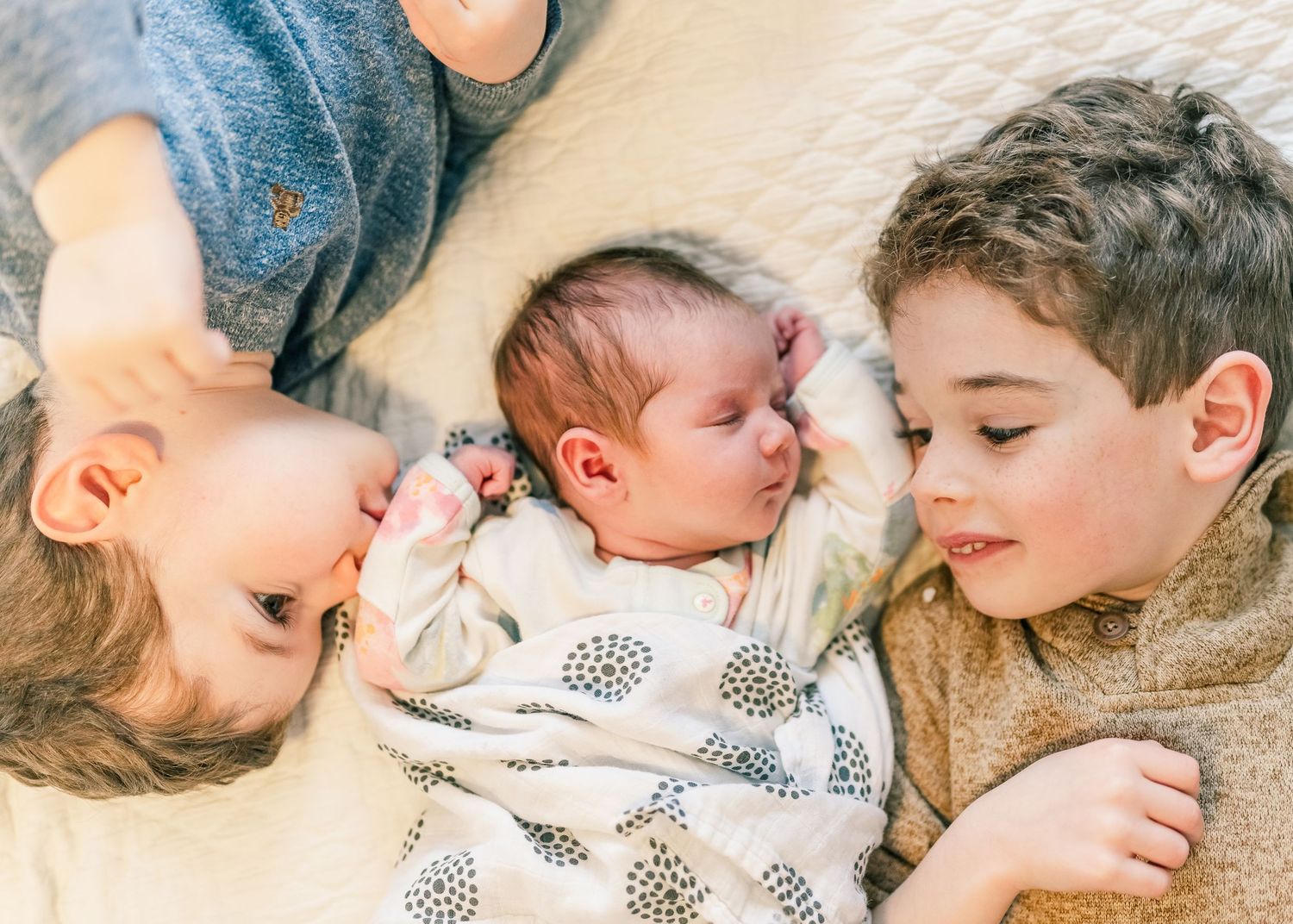Three siblings lying close together on white bedding, with the baby in the middle wearing a patterned outfit.