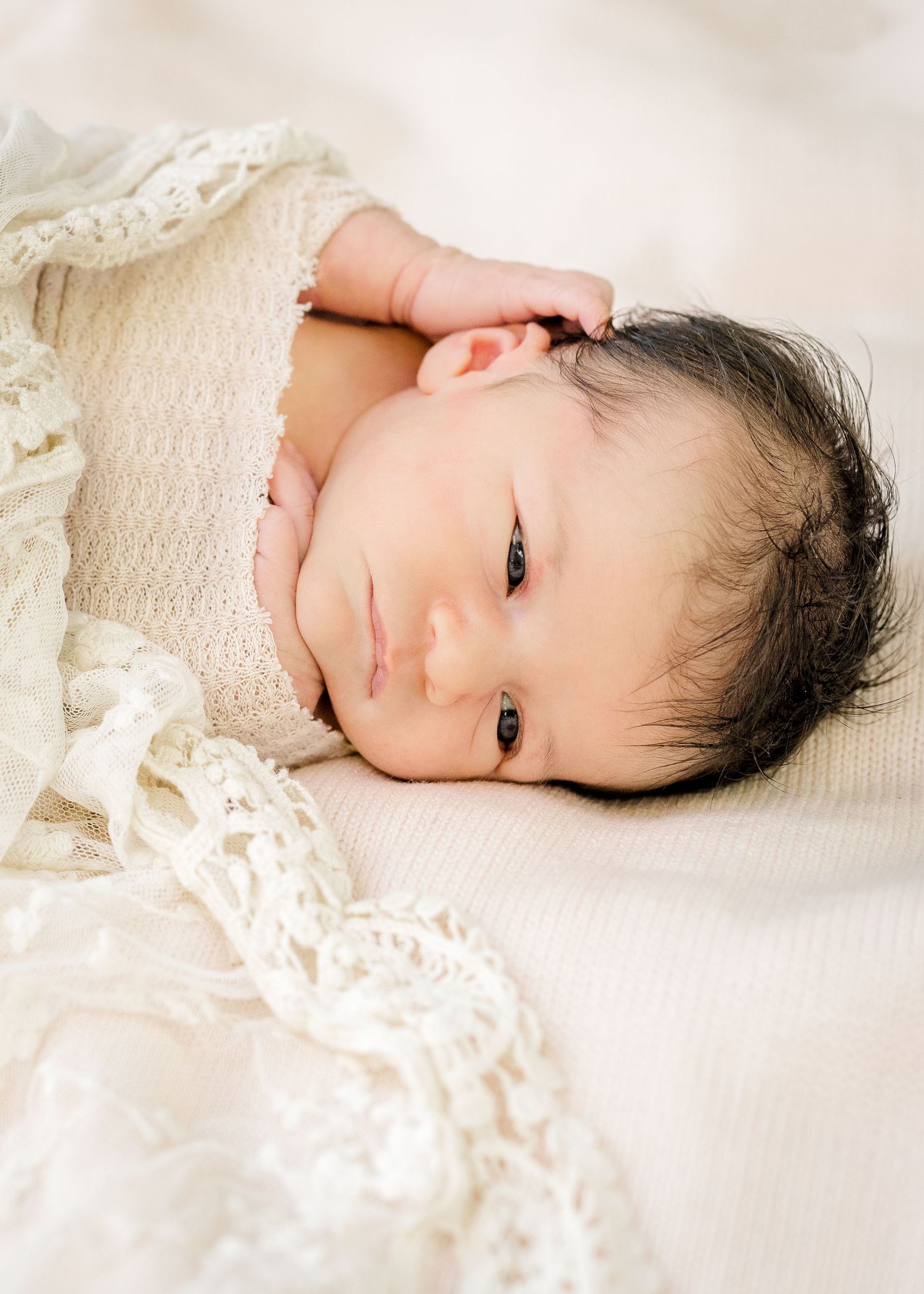 Peaceful newborn wrapped in cream lace blanket lying on white bedding.