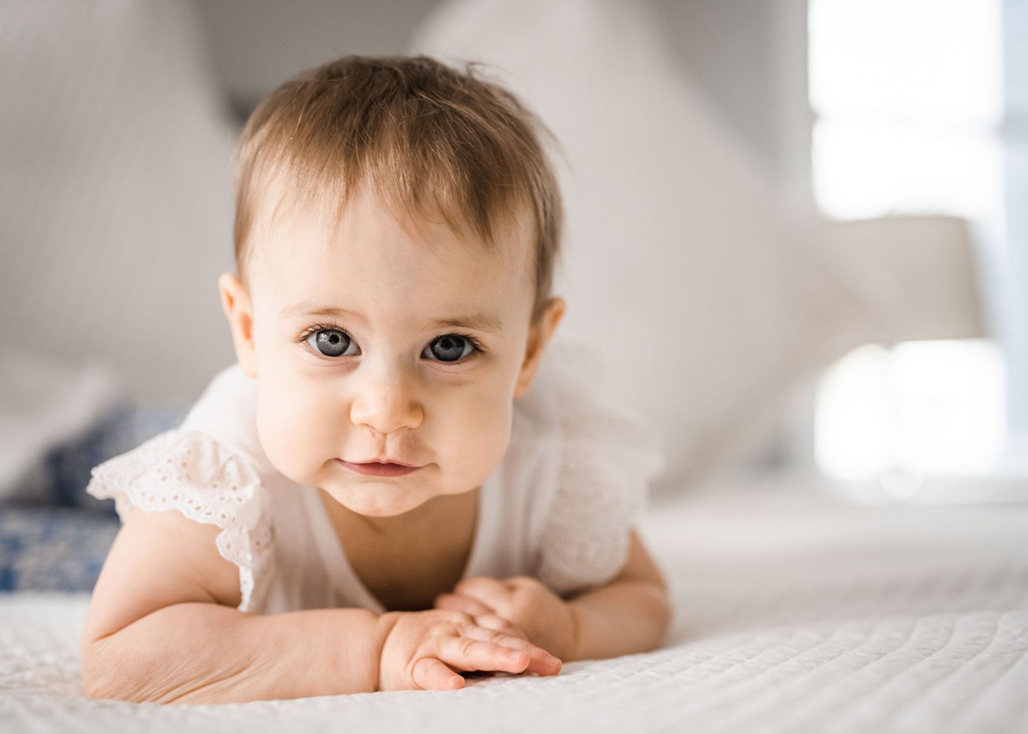 A baby in a white lace outfit lying on their tummy on white bedding.