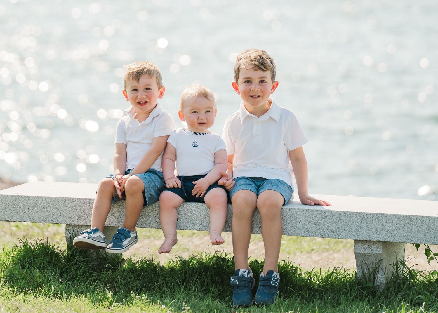 Three children sit on a white bench by the water's edge on a sunny day with sparkling bokeh in the background.