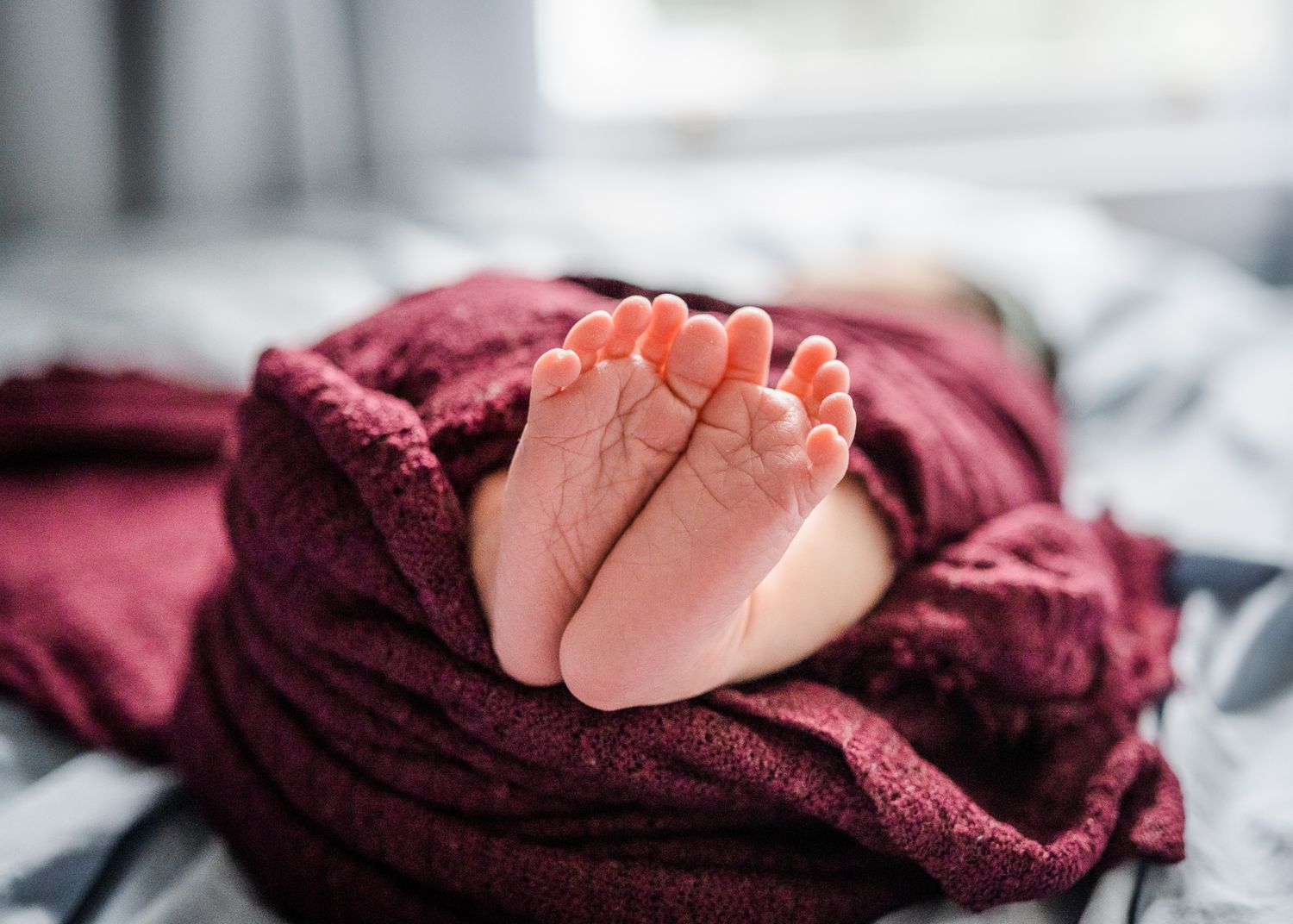 Close up of tiny newborn baby feet against burgundy blanket in natural window light.