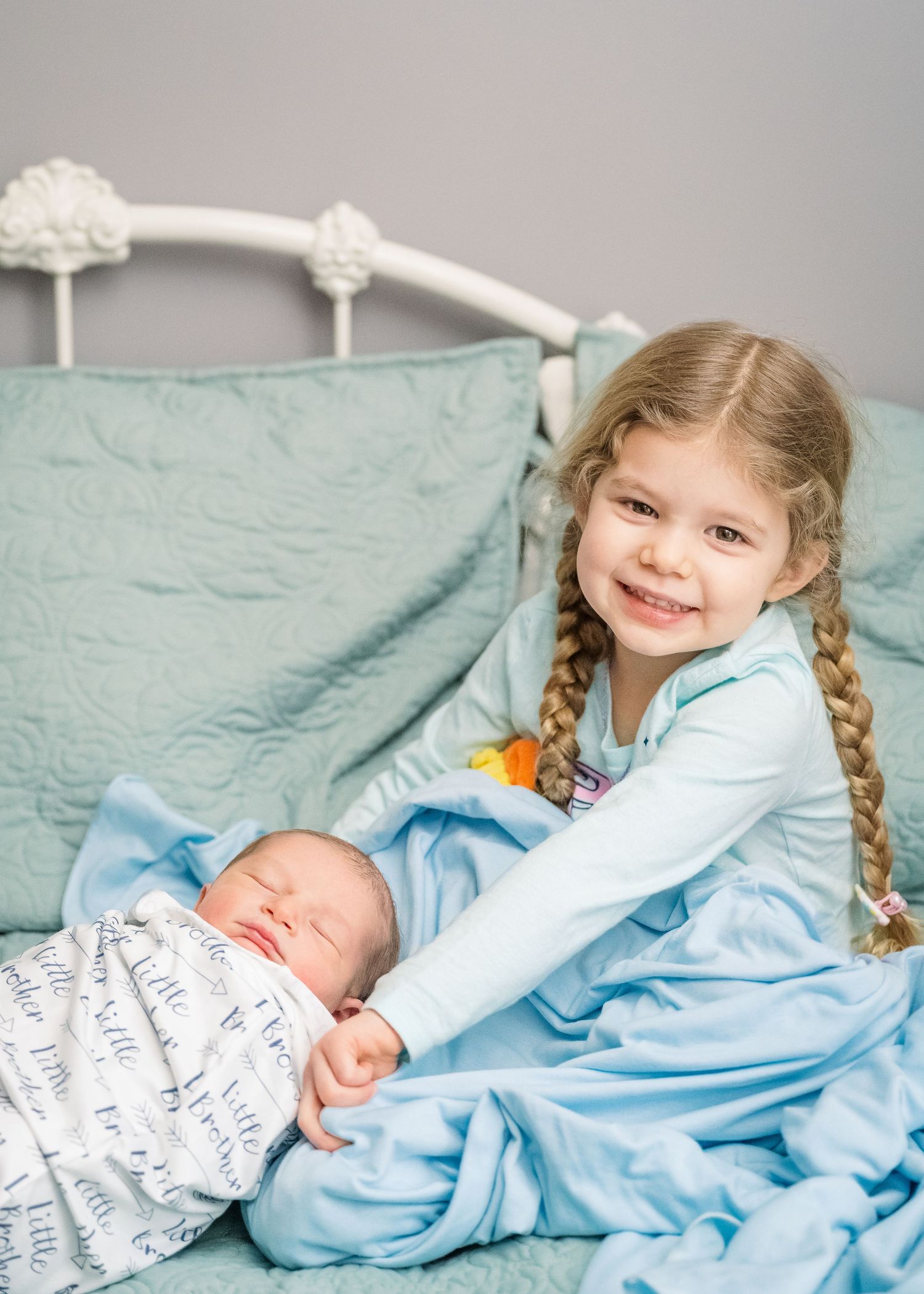 A joyful sibling moment on a bed with a newborn baby wrapped in a patterned blanket.