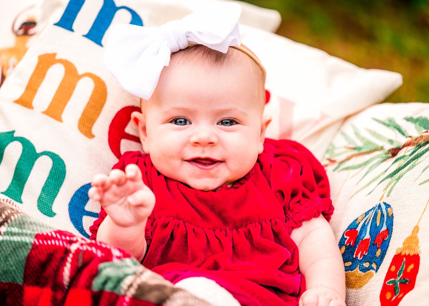 Baby in red dress smiles while laying on colorful holiday pillows.