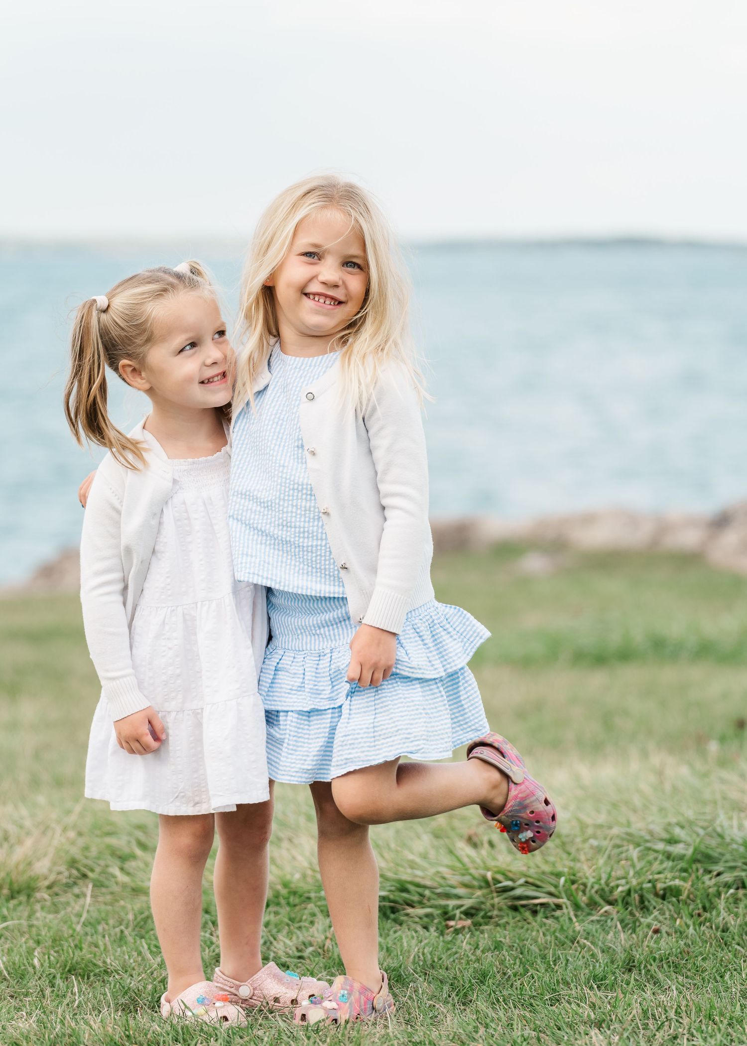 Two young children in white and blue dresses laugh and play together on grassy seaside hill.