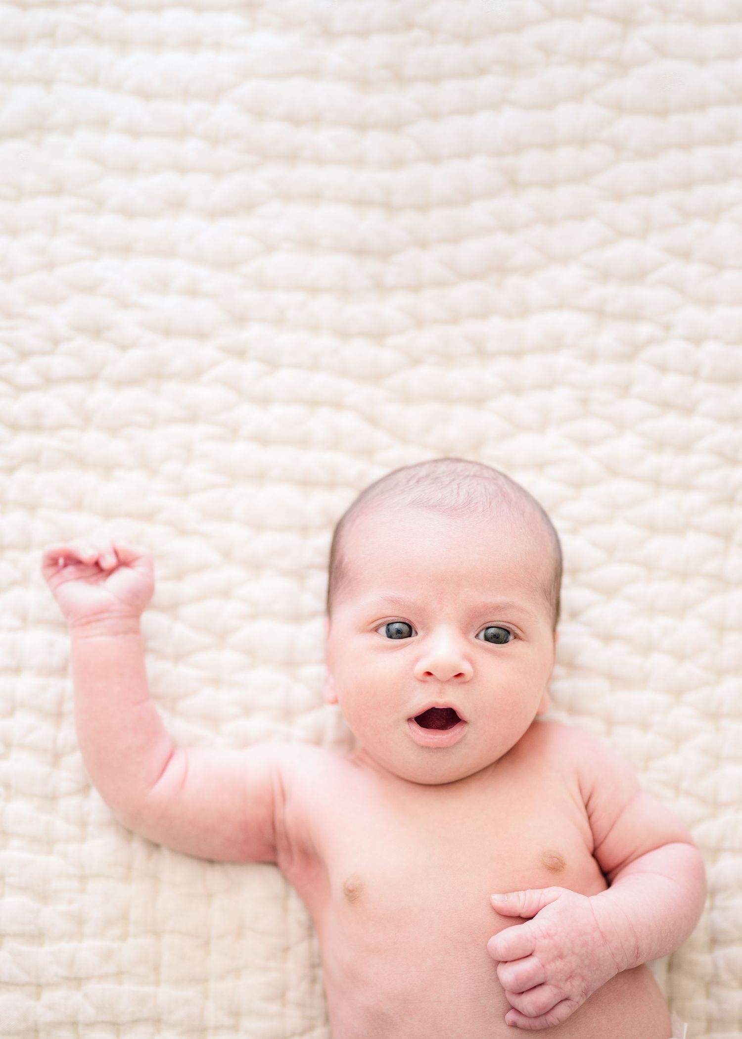Baby lying on white textured blanket with arm raised.