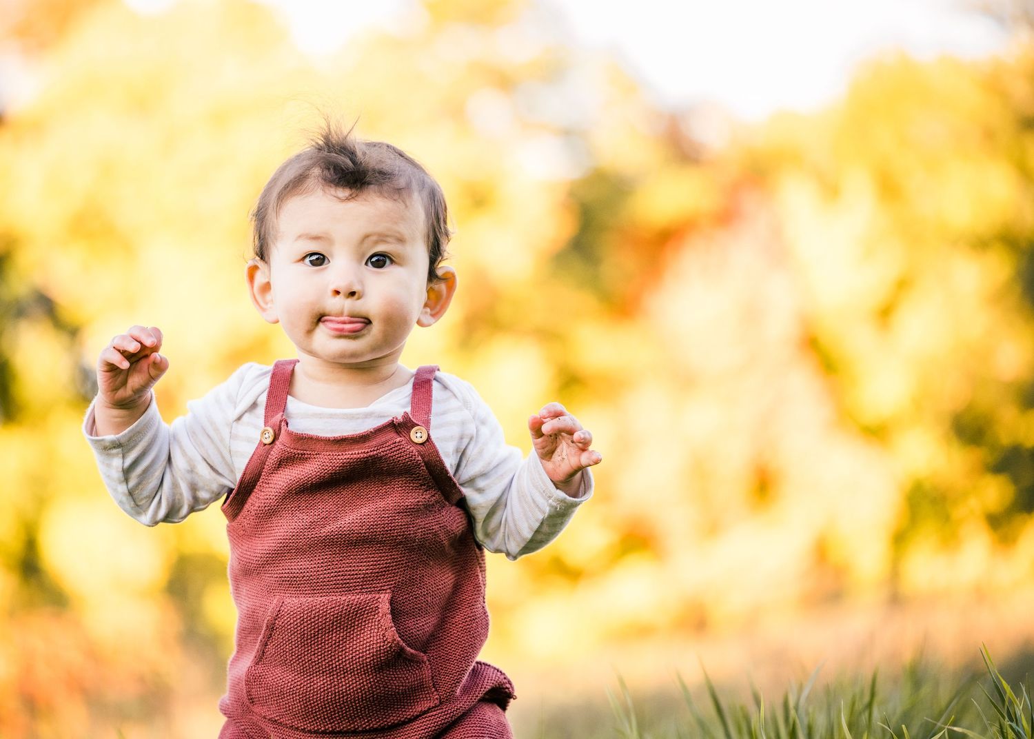 A cheerful baby in burgundy overalls enjoys playtime against vibrant autumn foliage.