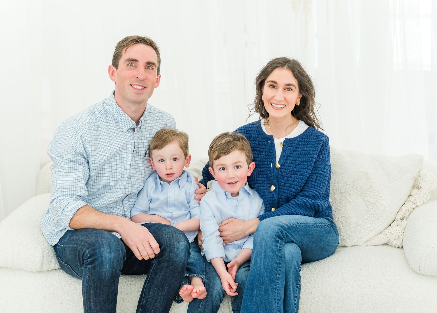 A family of four sits together on a white couch wearing casual blue outfits for a portrait session.