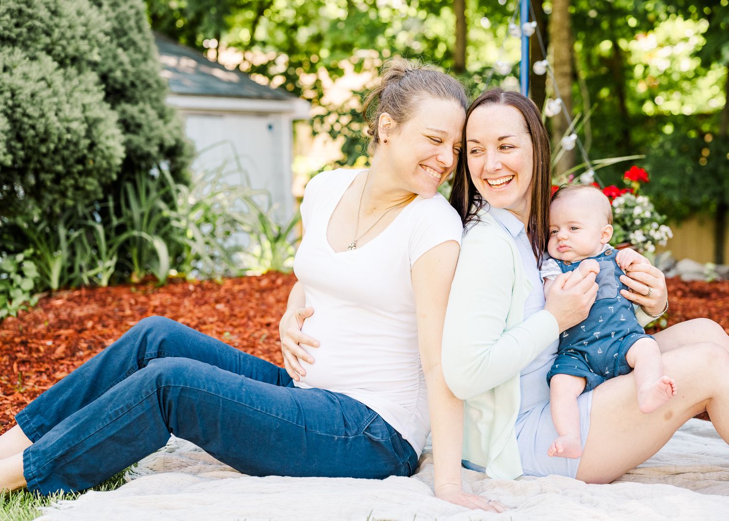 Family relaxes together outdoors on white blanket with mulch garden background.