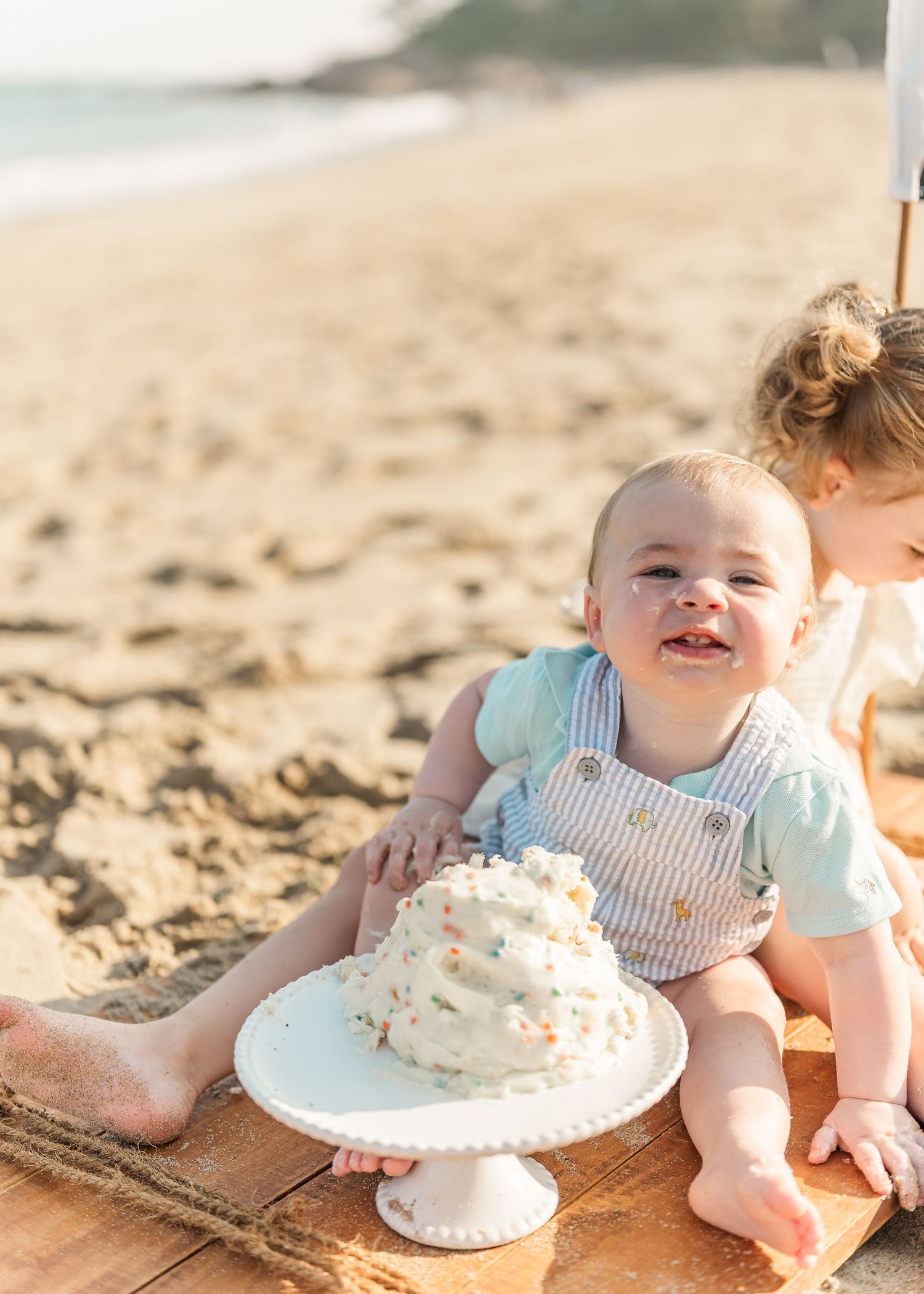 Baby in white outfit sits on beach blanket with birthday cake smash sequence showing messy frosting fun.