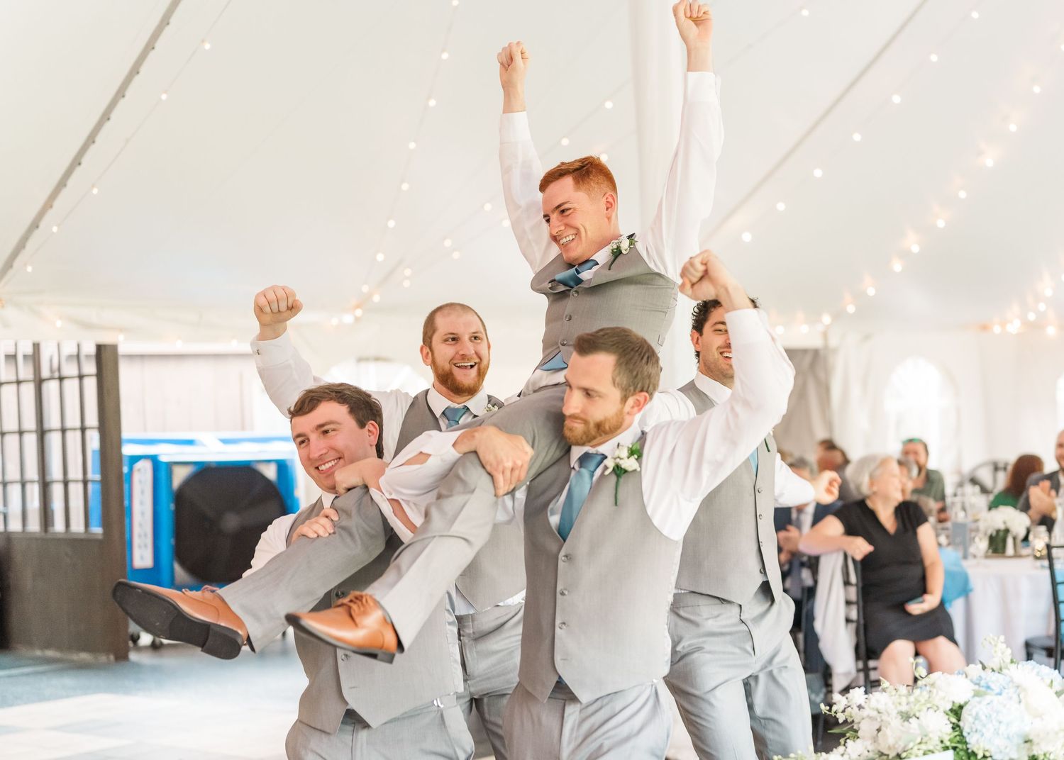 Wedding party in gray suits dancing in a sequence of celebratory poses under a white tent.