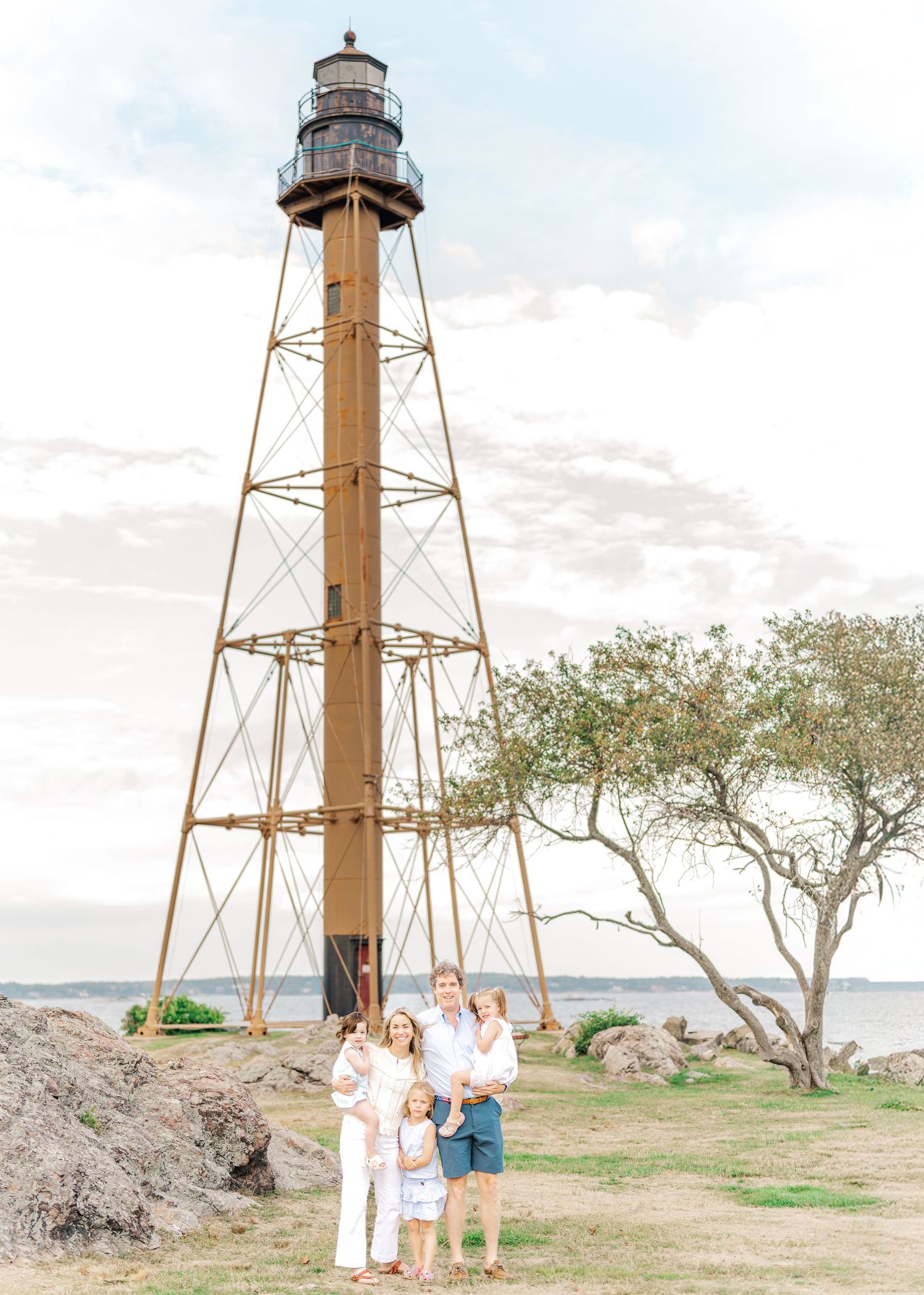 A tall rustic lighthouse tower stands on grassy coastal terrain with a tree and people gathered near its base.
