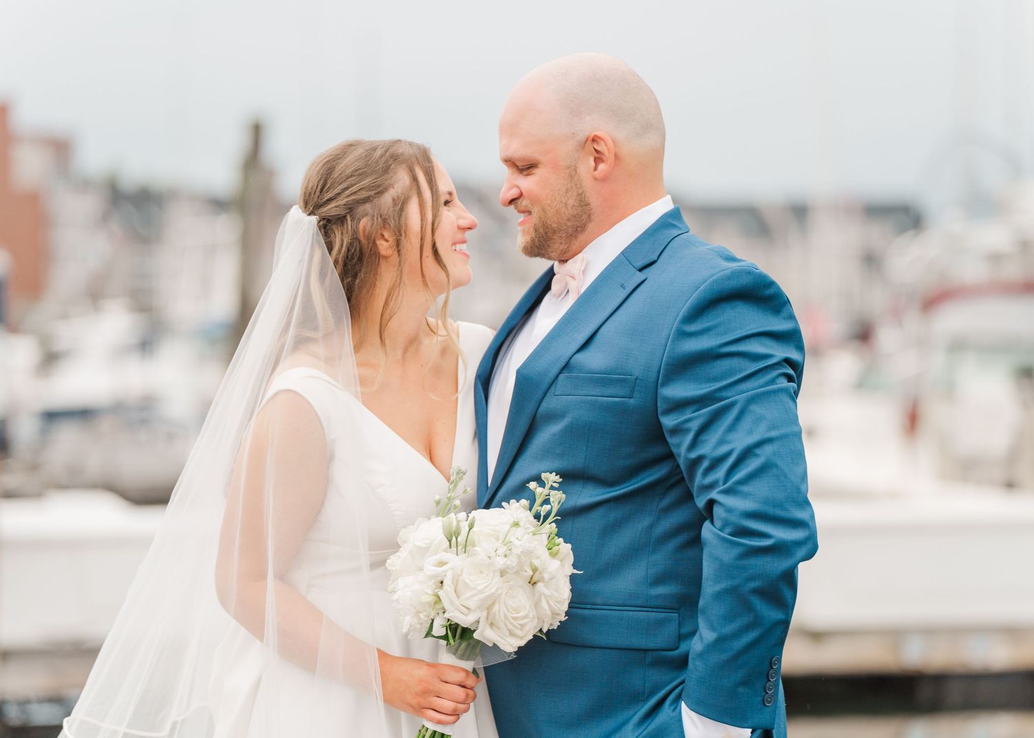 A couple in wedding attire embrace at a marina, with the bride holding a white bouquet and groom wearing a blue suit.