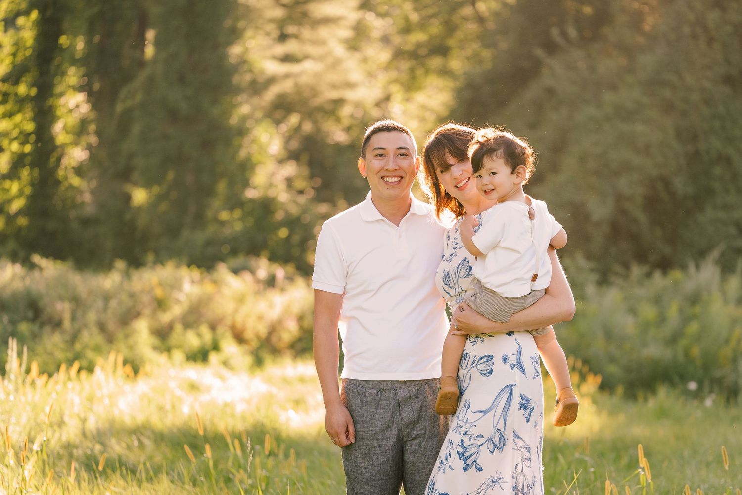 A sweet family portrait taken outdoors in warm evening sunlight with natural greenery in background.