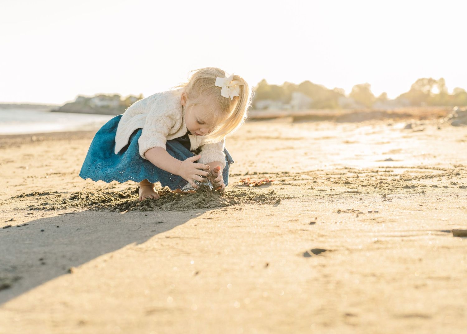 Person crouching on a sandy beach at sunset examining something on the ground.