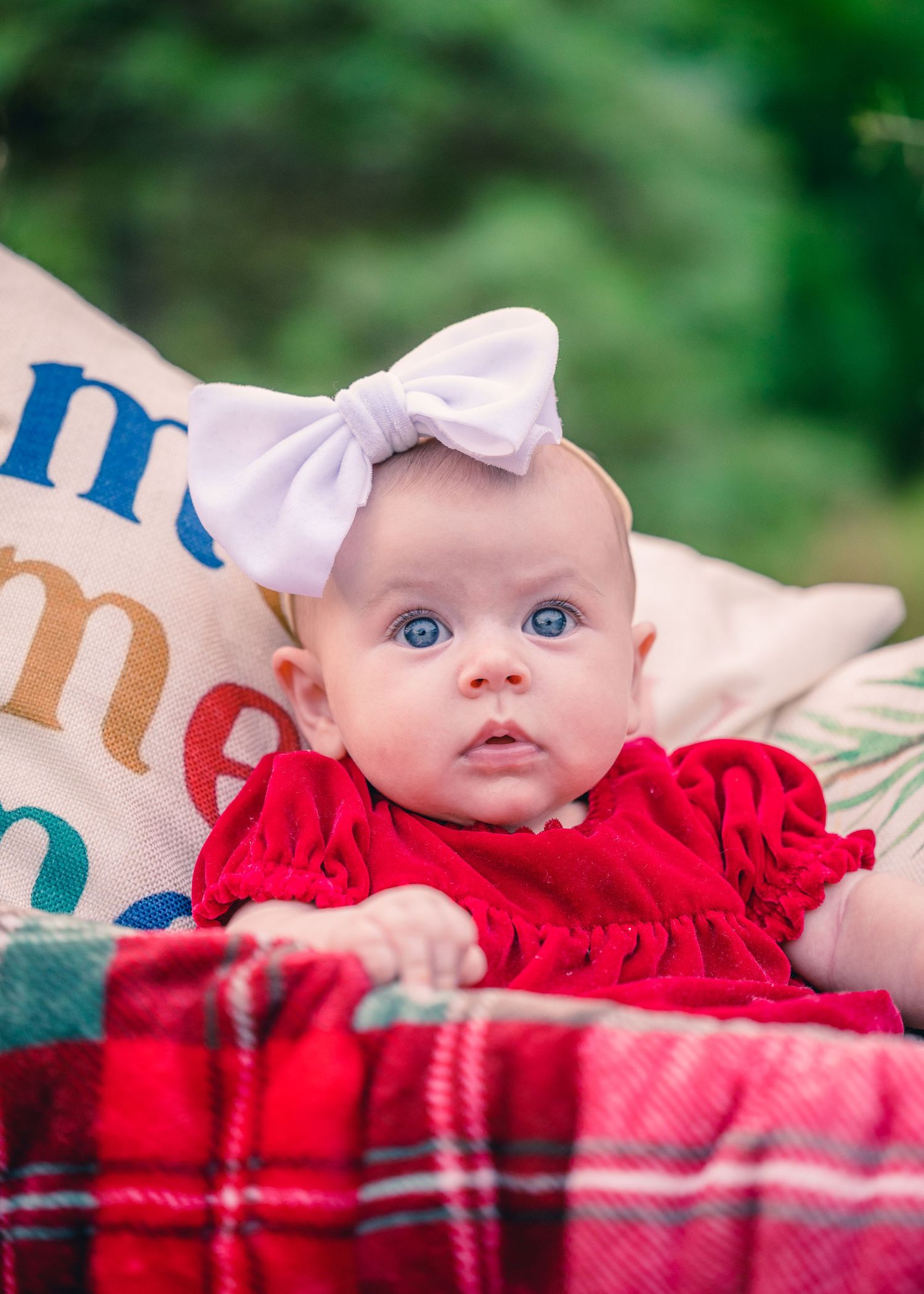 Baby in red dress with white bow headband lying on plaid blanket with decorative text pillows.