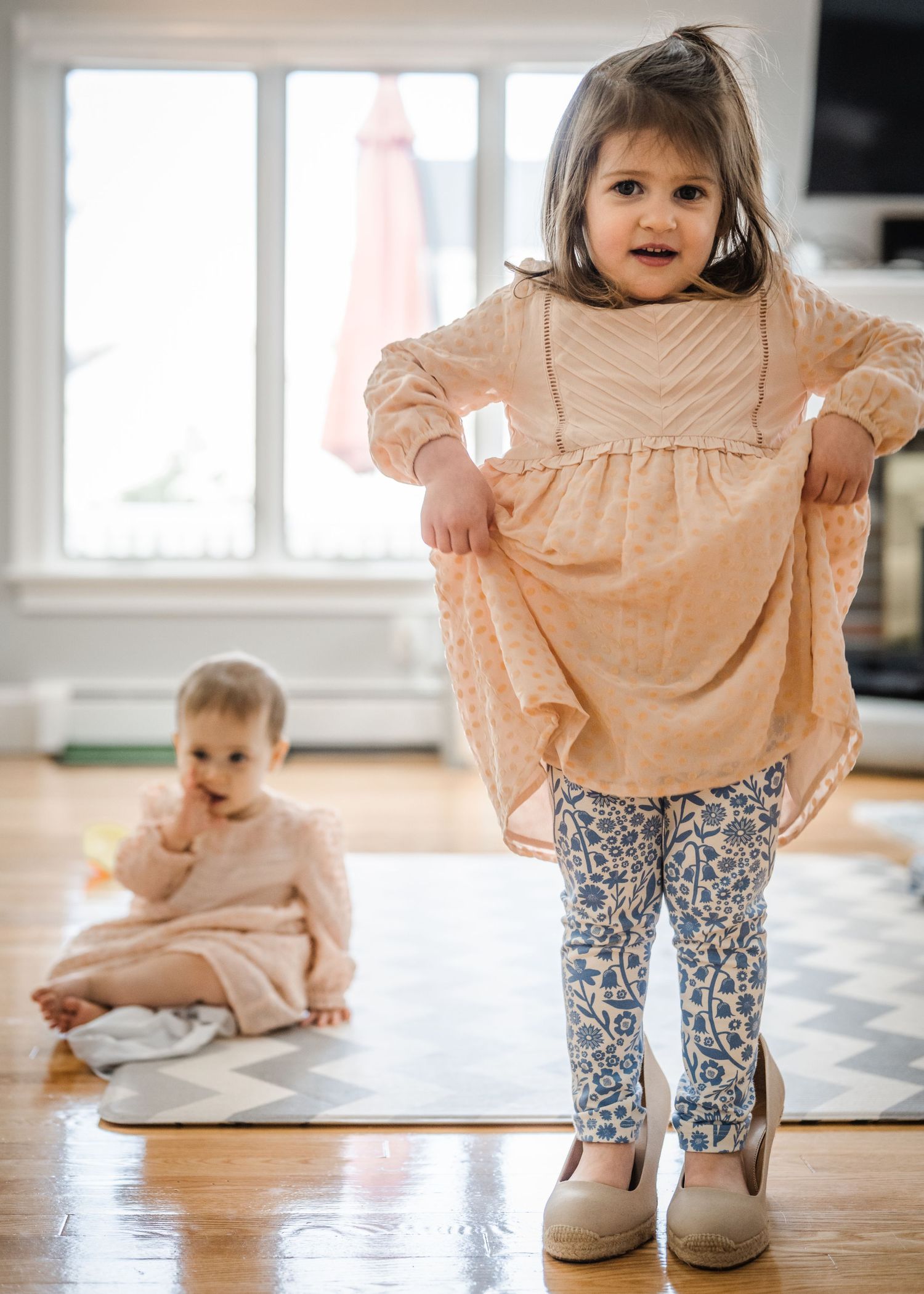 A child in a peach dress poses while another sits on the floor in a bright room.