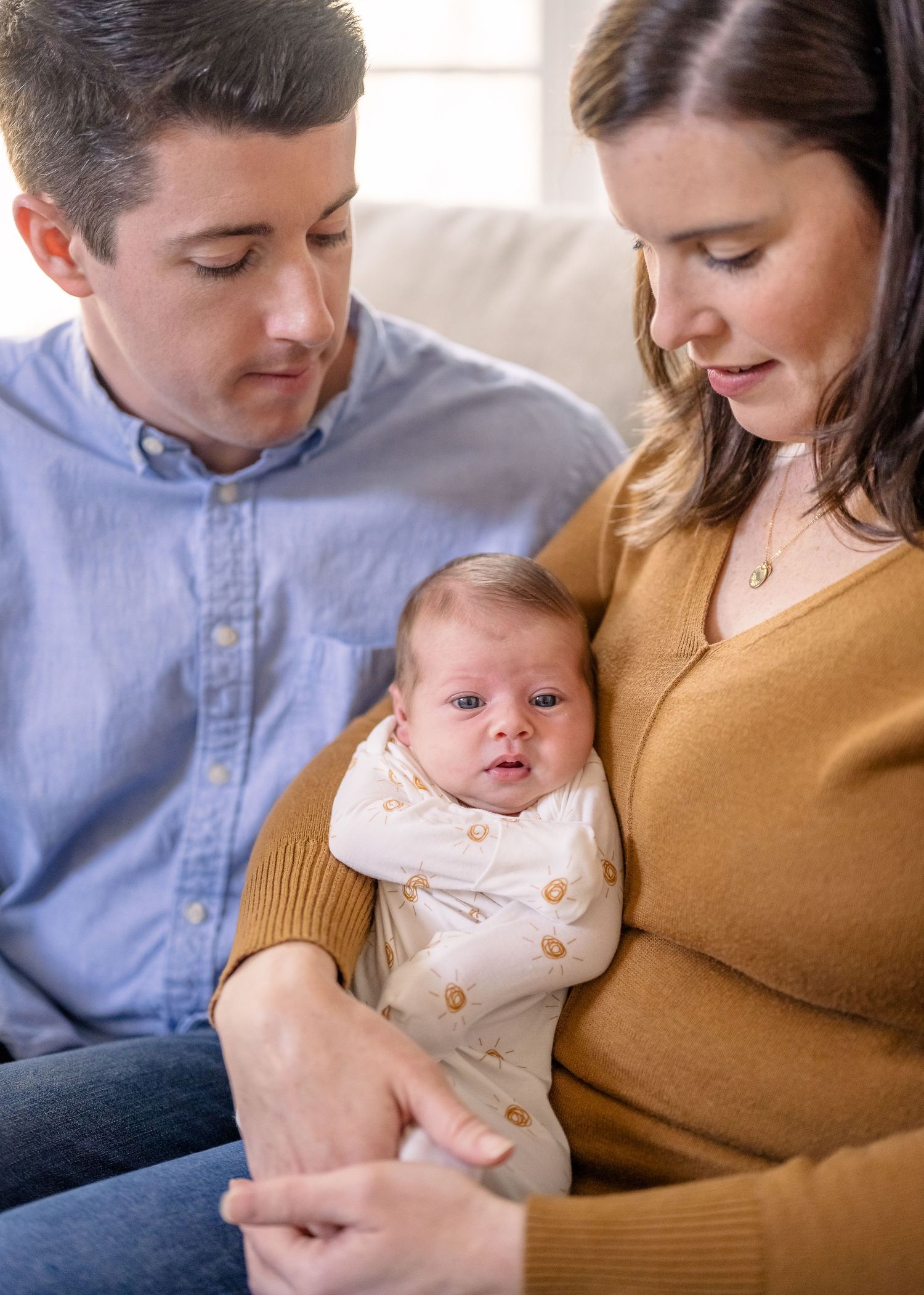 A tender family moment as parents bond with their infant wearing a white onesie on the couch.