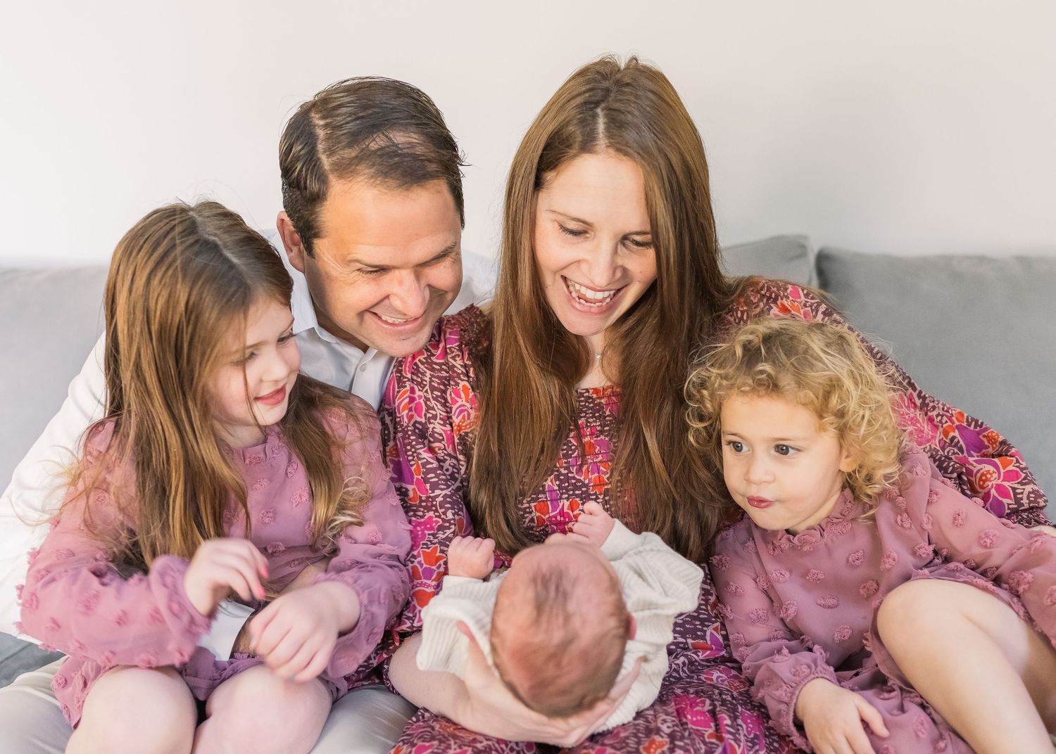 Family gathered on grey couch sharing intimate moment with new baby in coordinated pink outfits.