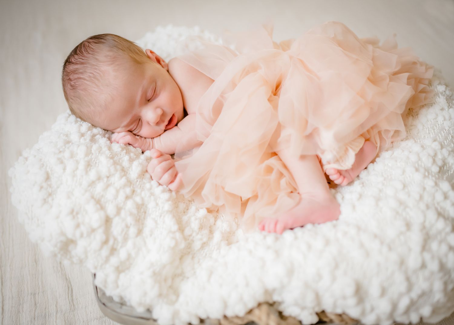 Sleeping newborn in peachy tulle dress lying on soft white textured blanket.