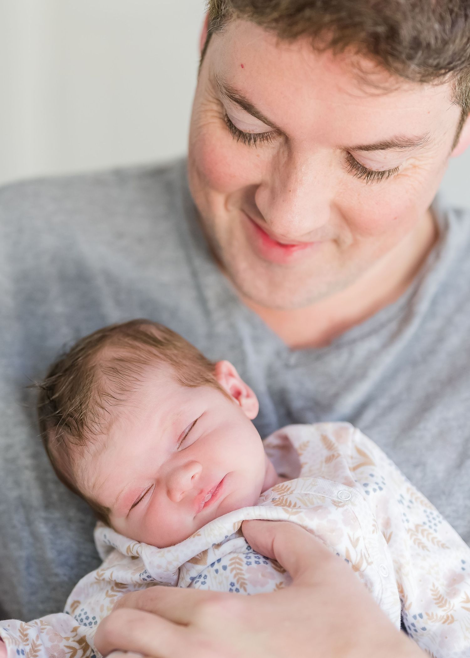 A parent wearing a grey shirt gently holds and gazes down at a sleeping newborn baby in white floral clothing.