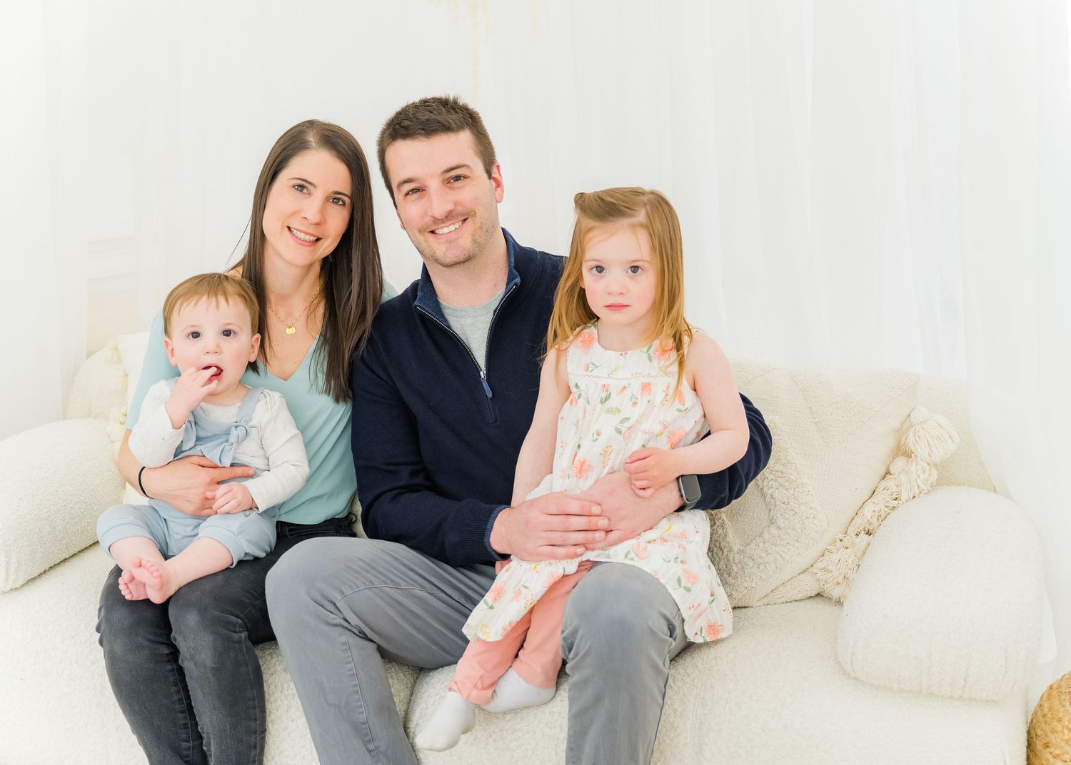 A family of four sits together on a white couch for a studio portrait wearing coordinated pastel colors.