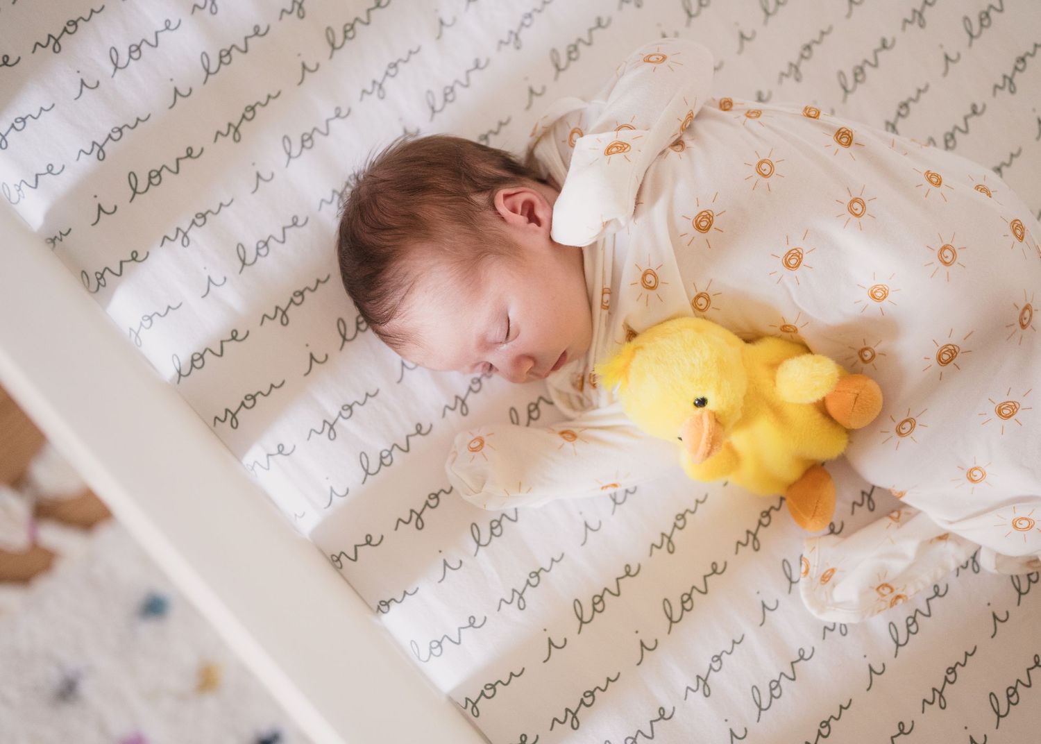 Sleeping baby in white onesie lies next to yellow plush toy on patterned bedding.