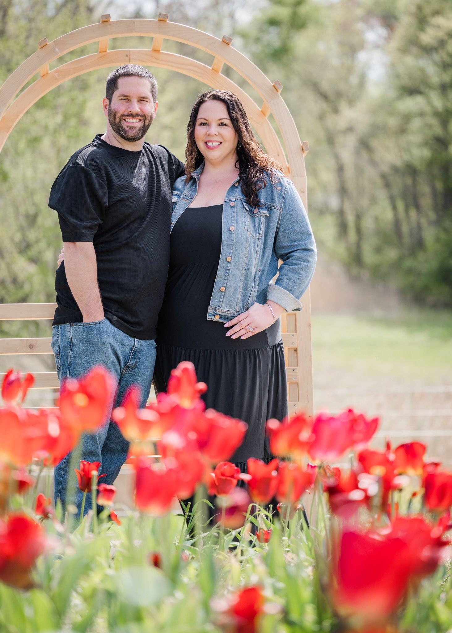 Couple standing together under a wooden garden arch with red tulips in foreground.