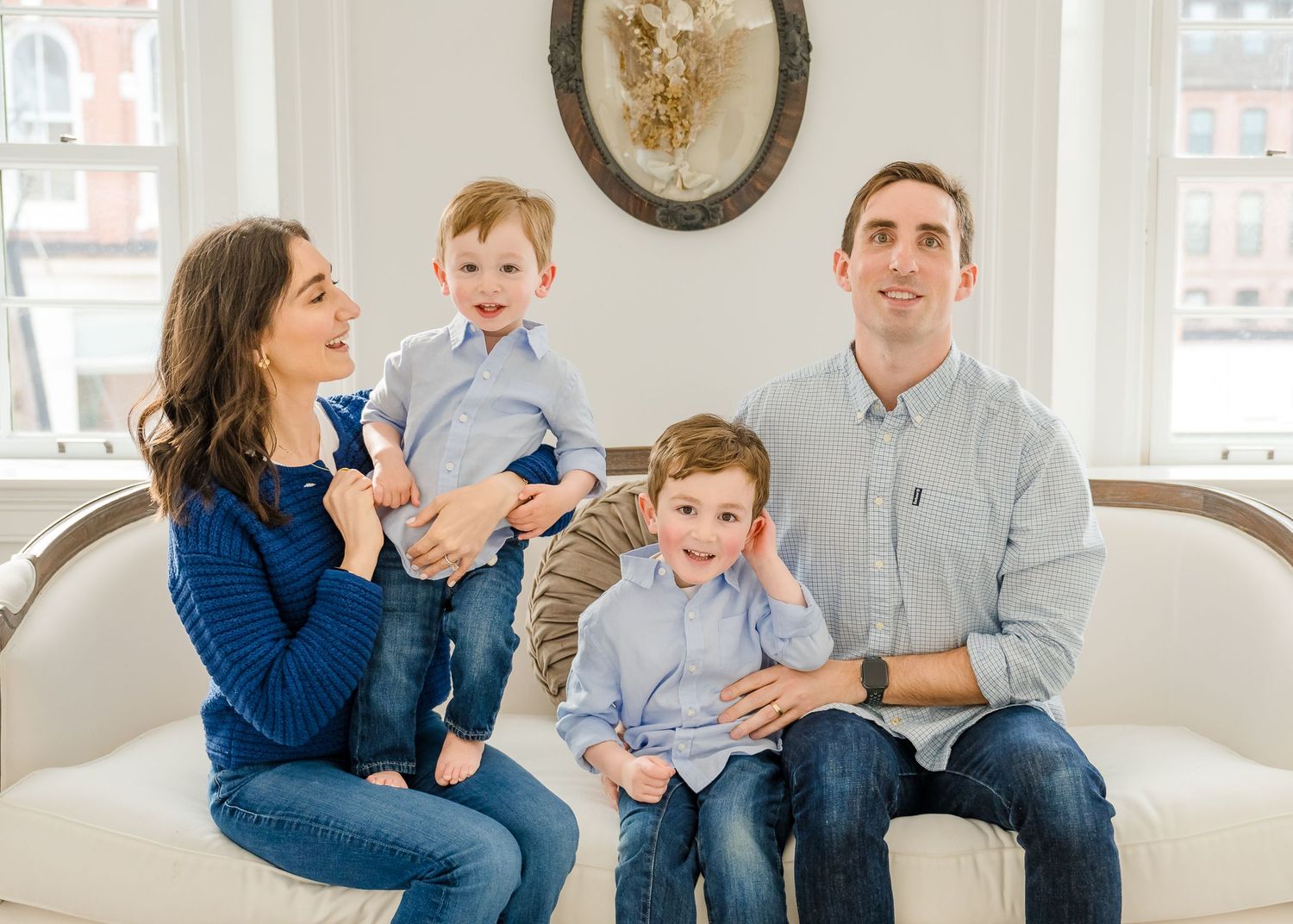 A family of four sits together on a light gray couch in a bright room with a round wall decoration.
