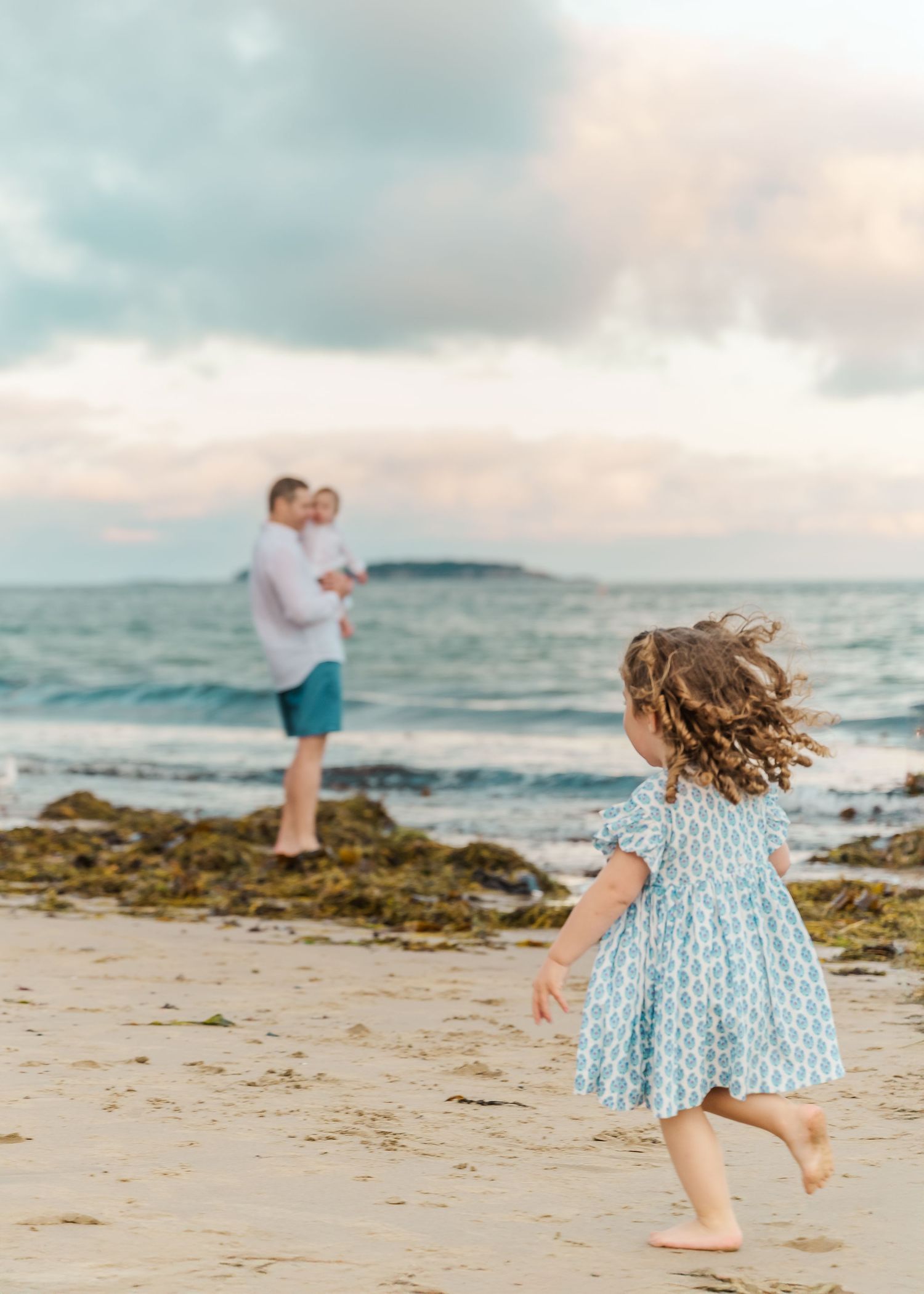 A young girl in a blue patterned dress twirls joyfully on a beach at sunset while others stand near the shore.