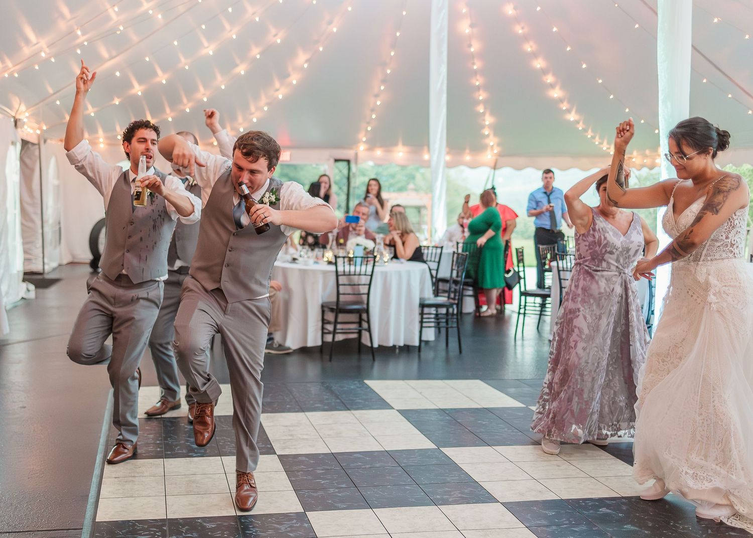 Wedding party members perform dance moves in a bright reception tent decorated with hanging string lights.