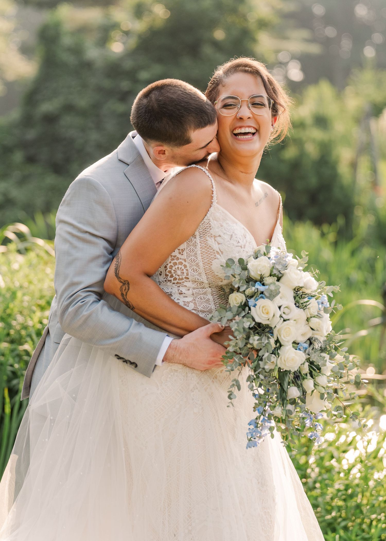 Joyful wedding couple embraces outdoors with white and blue floral bouquet in natural sunlit setting.