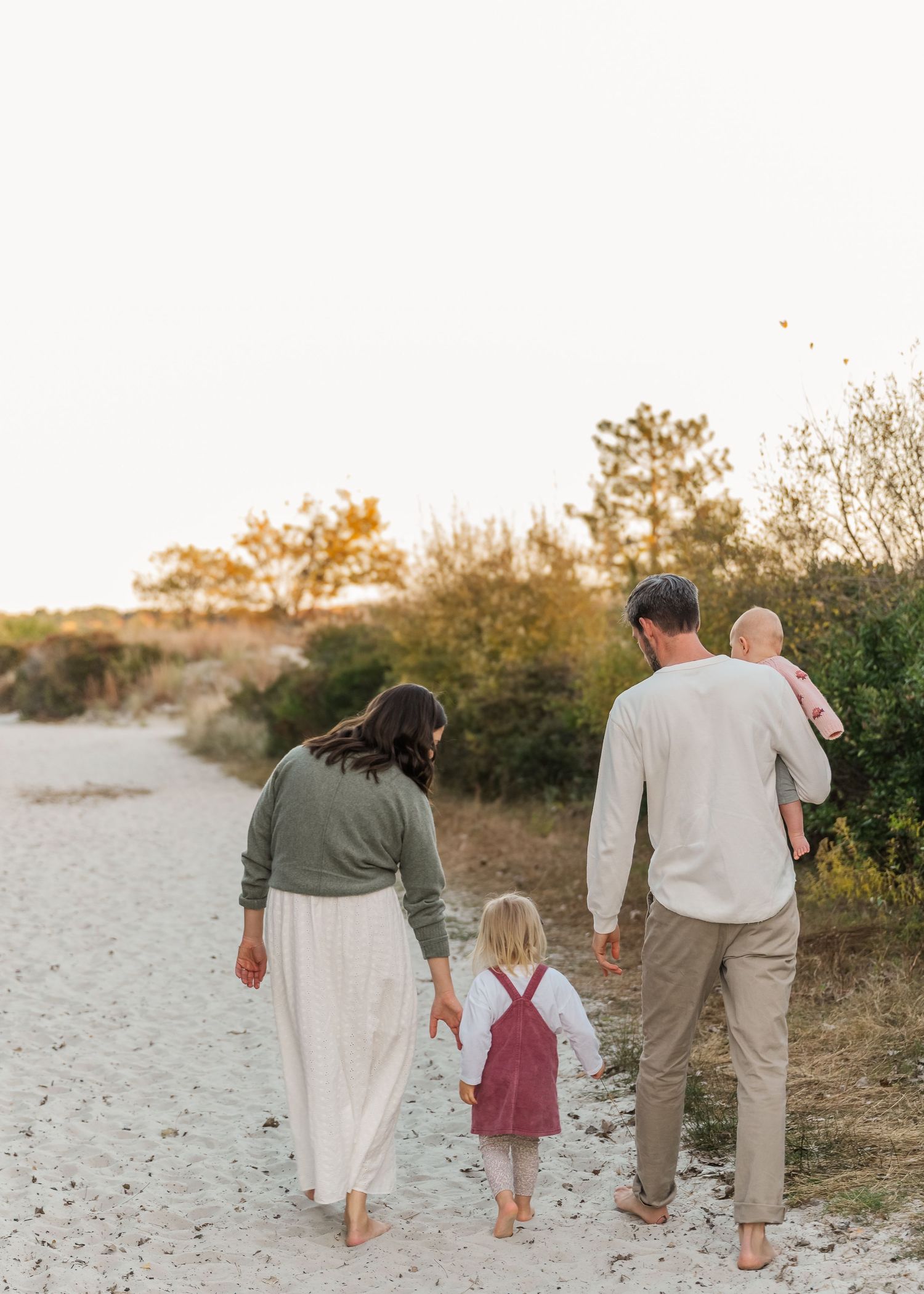 Family walking together barefoot on a sandy beach at sunset, holding hands in a line.