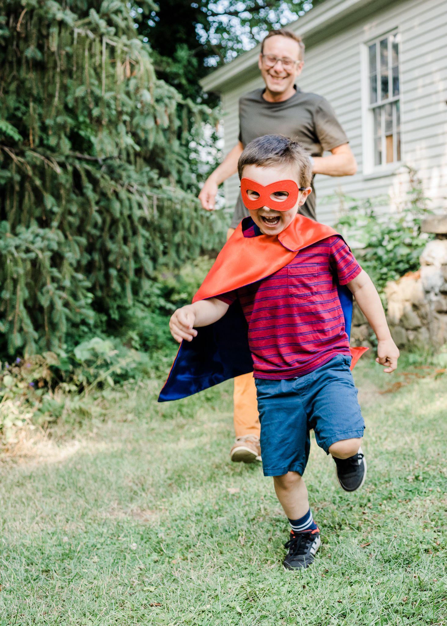 Child playing superhero with an orange mask and cape while running in a backyard.