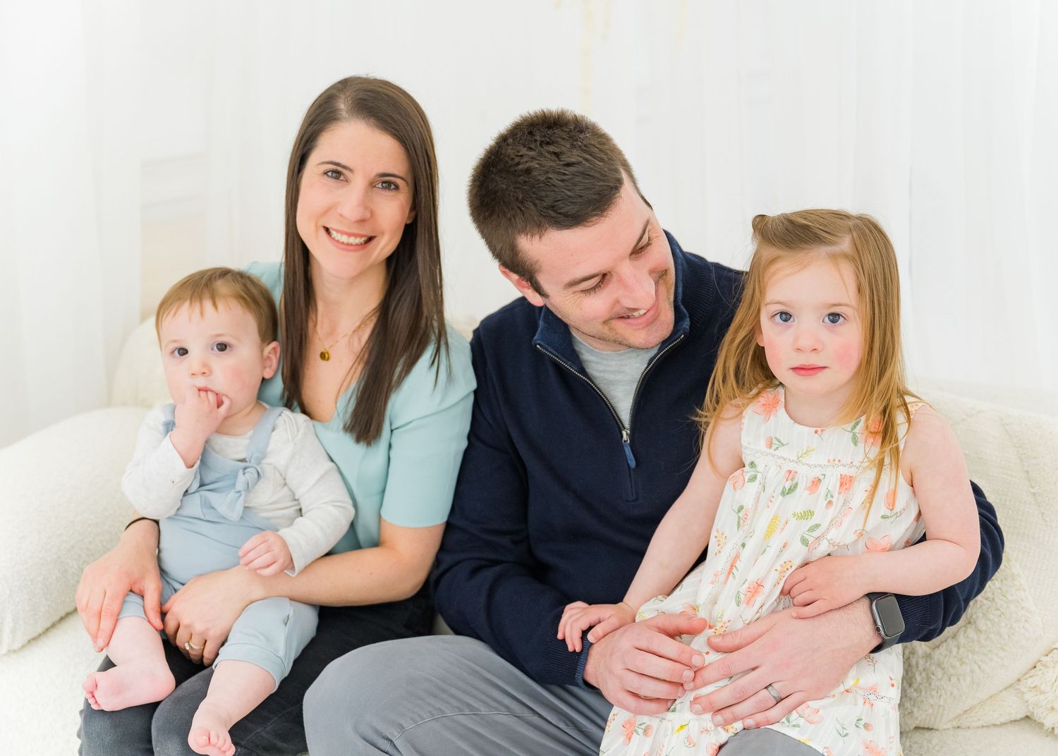 A family sits comfortably on a white couch, wearing casual attire in a minimalist studio setting.