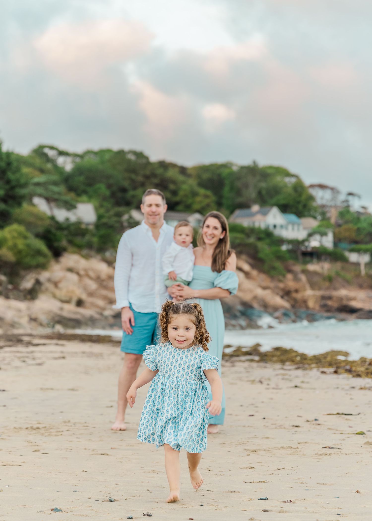 A family enjoys a relaxed moment together on a scenic beach with rocky coastline and houses on the hillside.