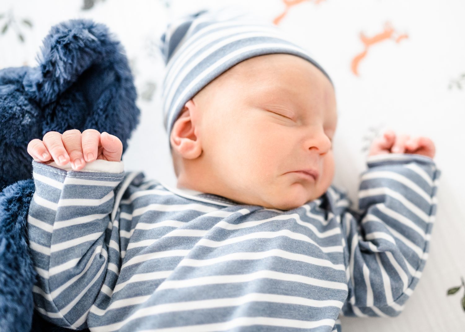 Sleeping newborn wearing grey and white striped outfit with arms raised on white bedding.