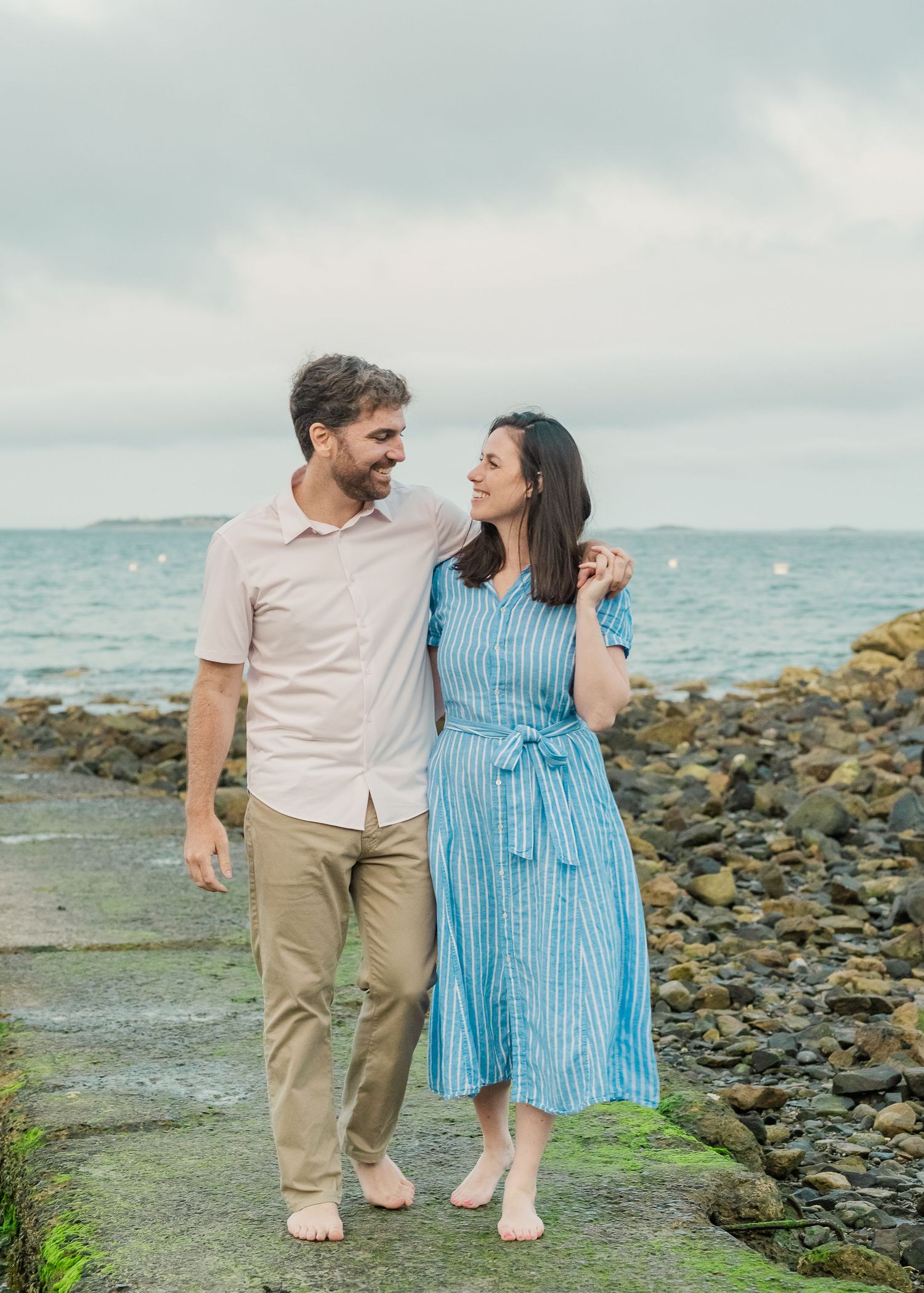 Couple walking barefoot along rocky coastline in casual summer outfits.