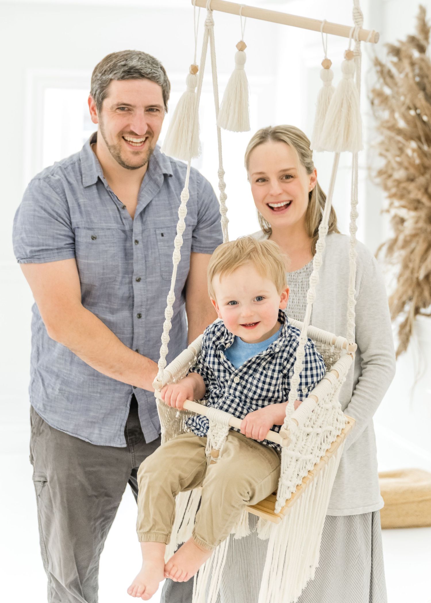 A smiling family poses together with a child on a macrame swing in a bright white room with boho decor.