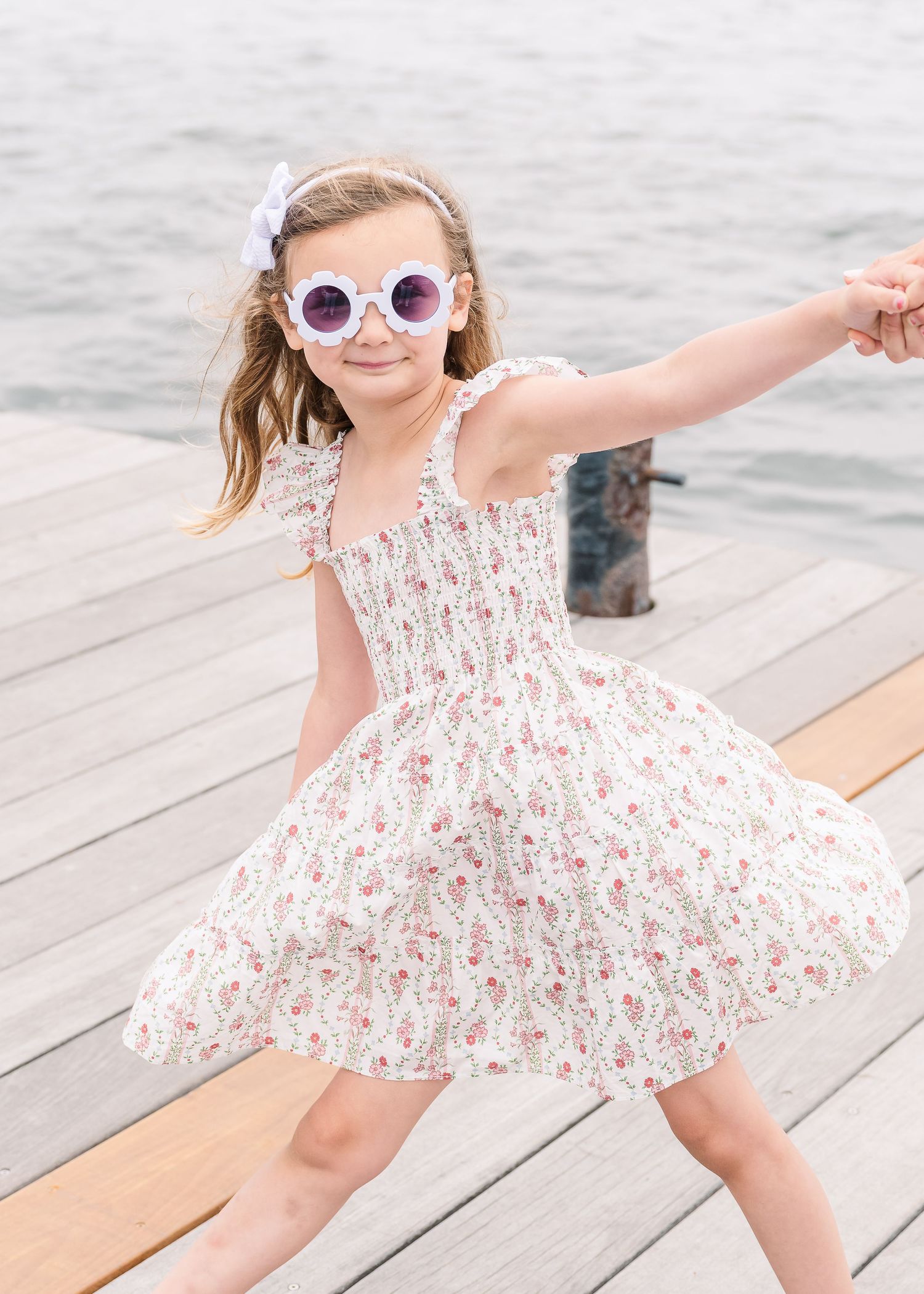 Young girl in a floral dress and sunglasses twirls on a wooden dock by the ocean.
